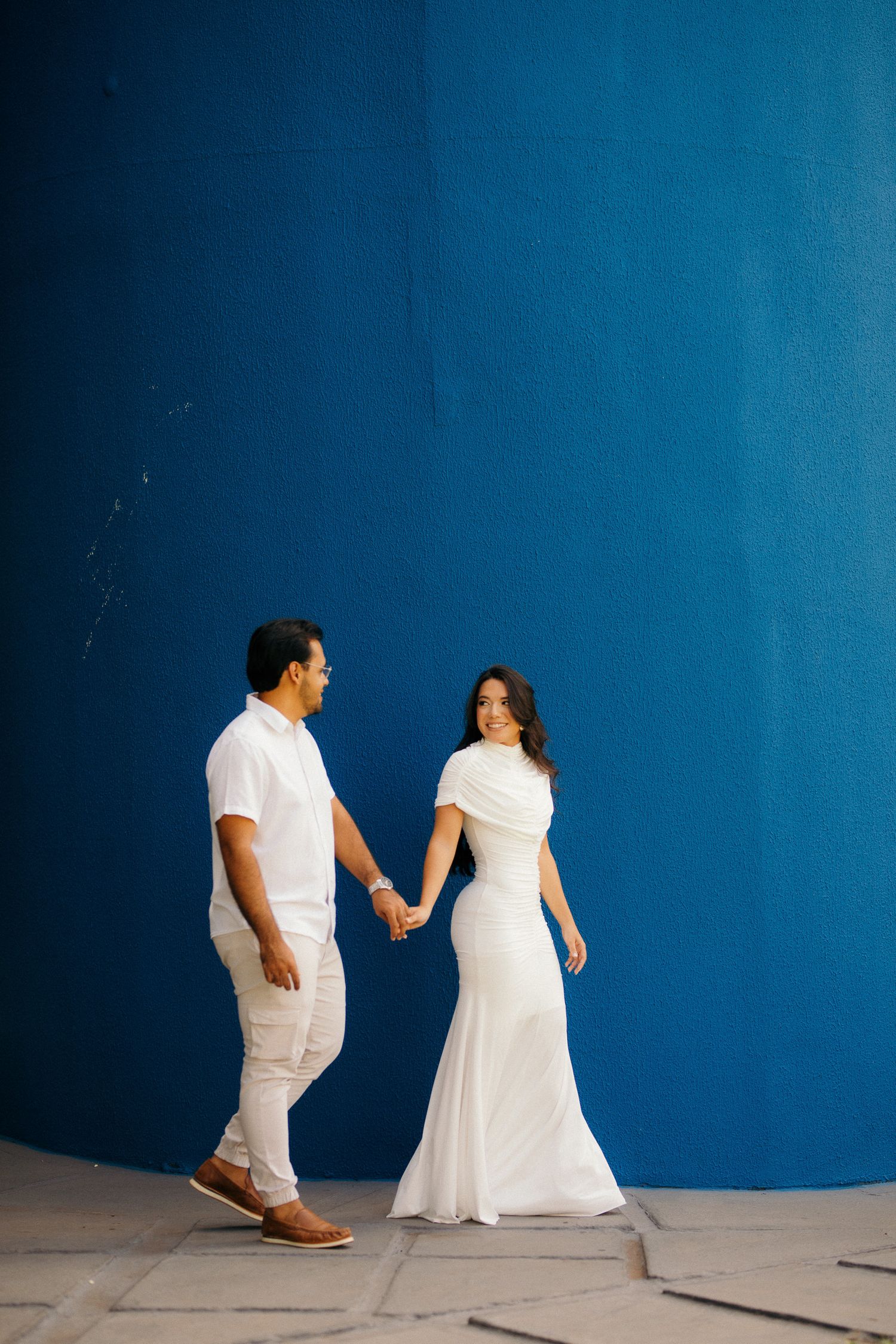 Couple holding hands in white outfits against a vibrant blue wall.