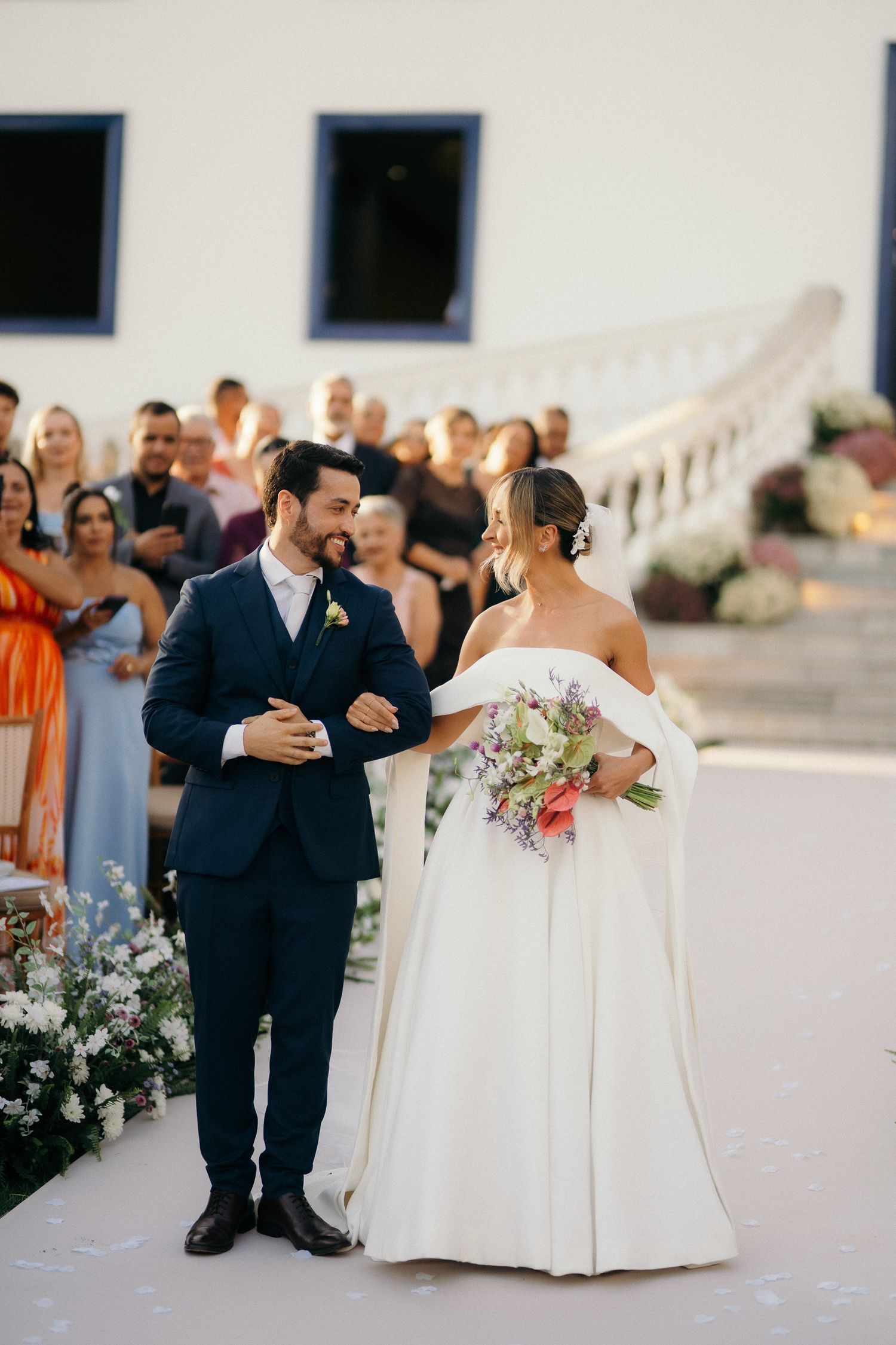 Bride and groom walk down the aisle together, bride holding colorful bouquet in elegant off-shoulder white gown.