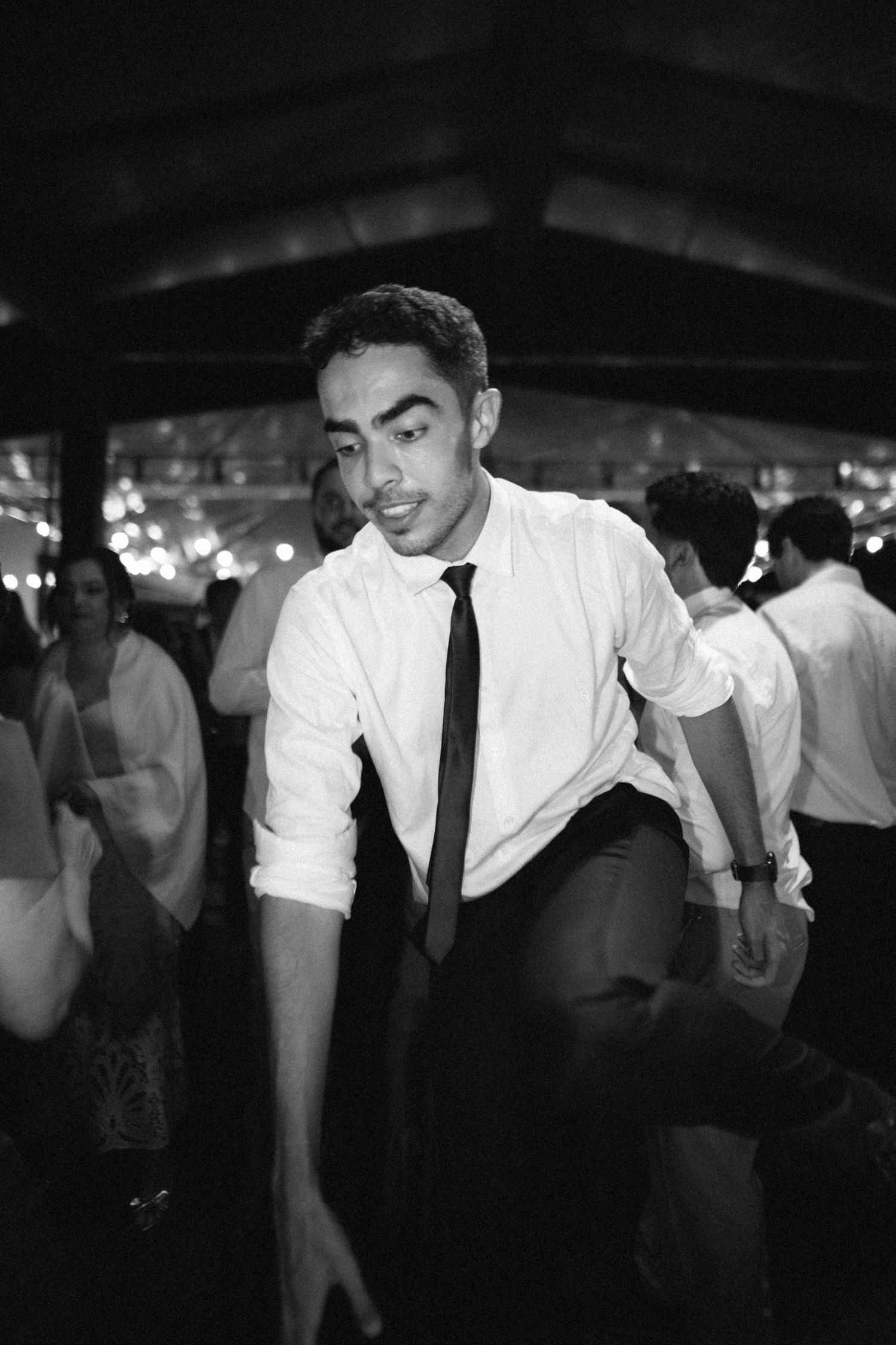 Black and white photo of a young man in a white shirt and tie dancing energetically at a wedding reception.