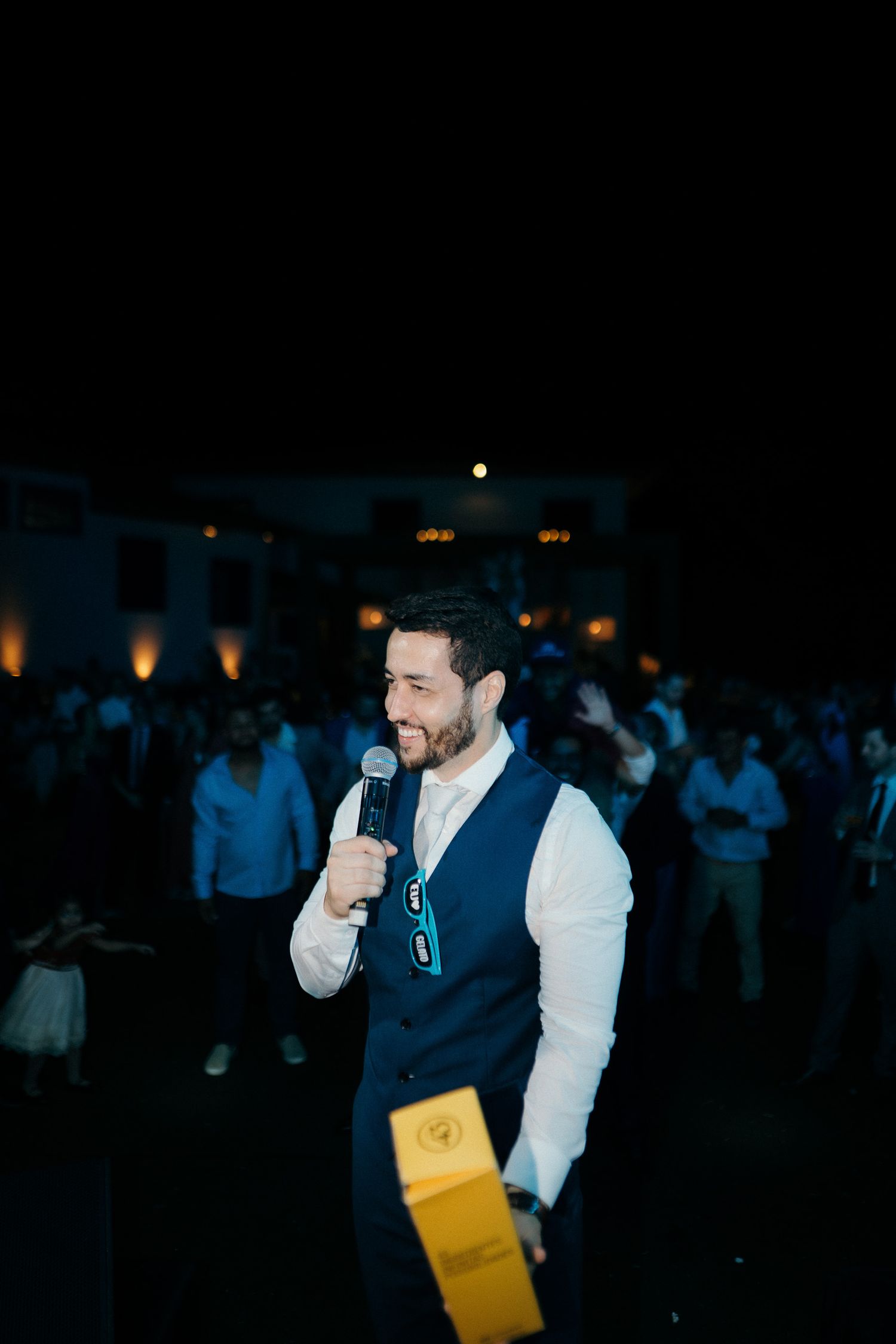 Smiling man in a navy vest speaks into a microphone at an outdoor evening event.