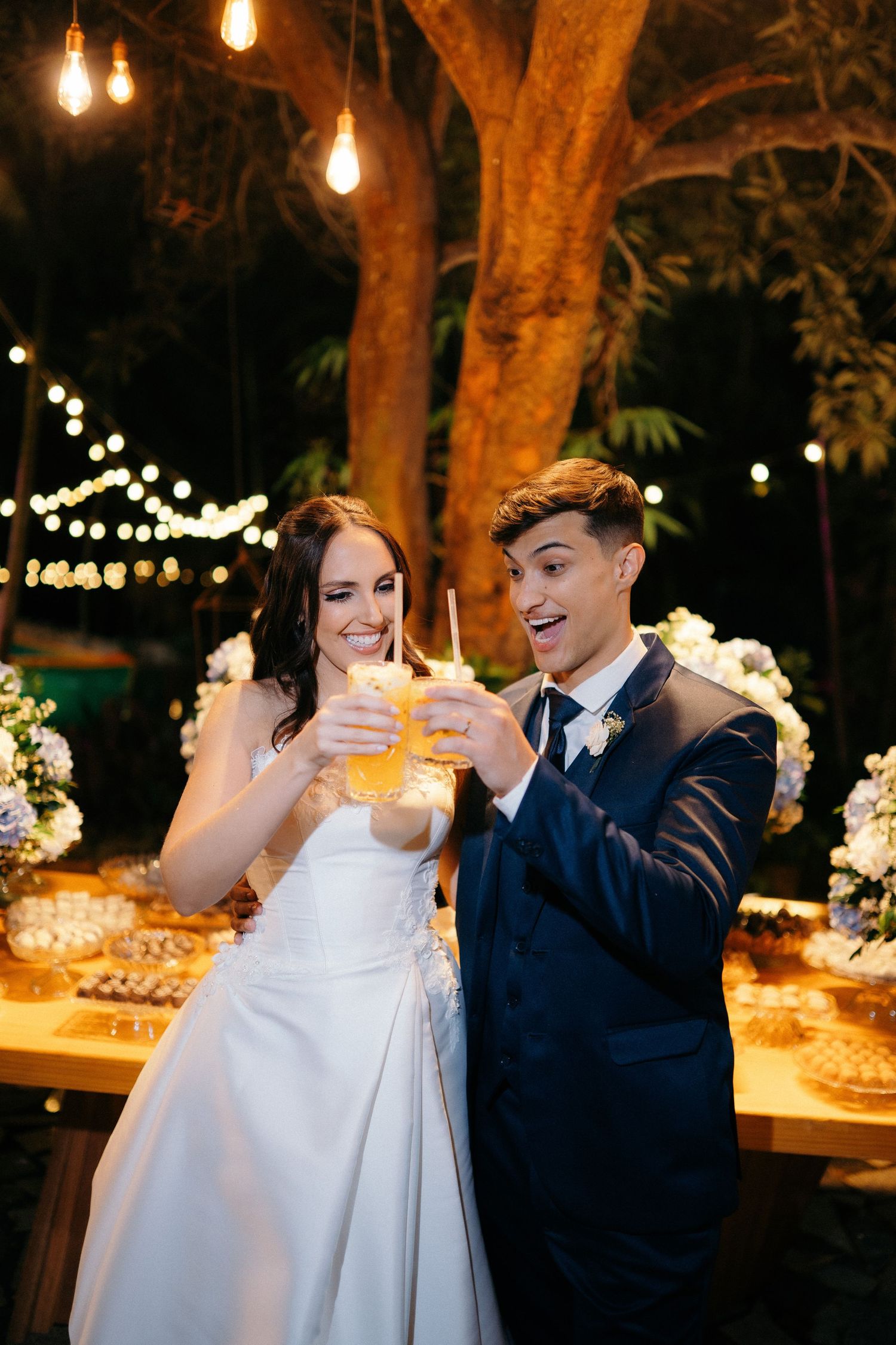 Smiling bride and groom toasting with drinks at their outdoor evening wedding reception under warm string lights.