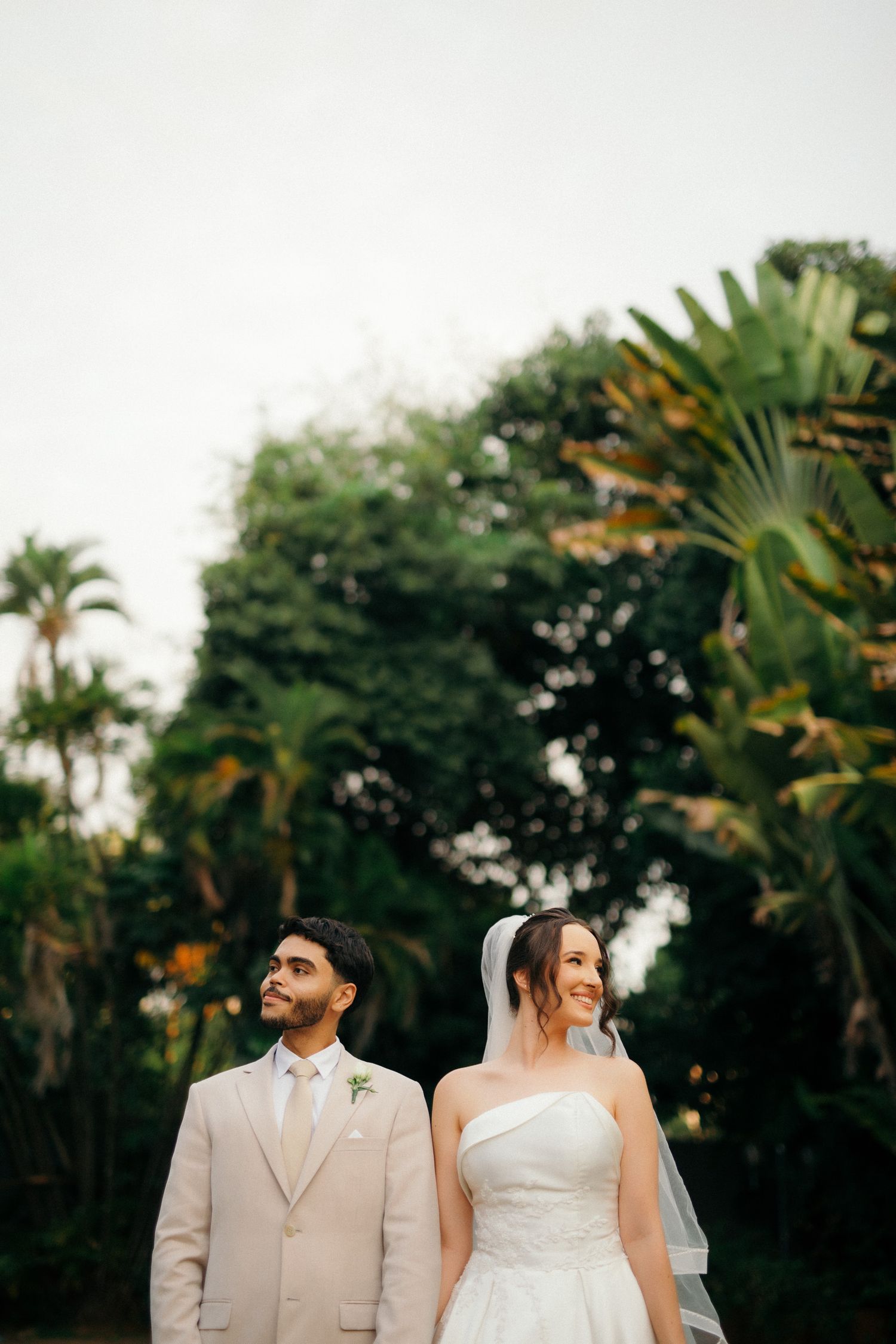 Bride and groom standing together outdoors with lush tropical greenery backdrop on their wedding day.