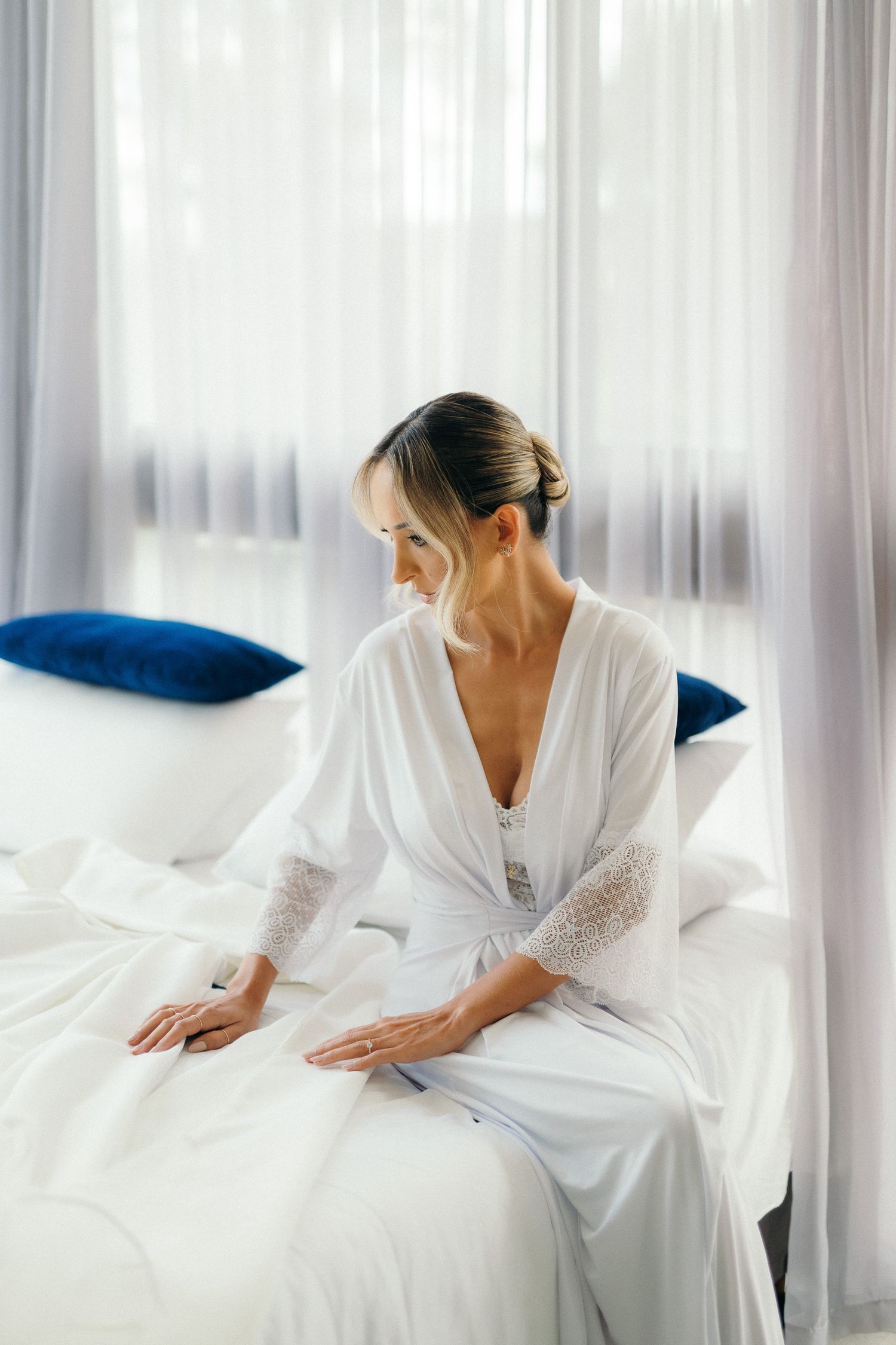 Elegant bride in a white lace robe sitting gracefully on a bed near sheer curtains.