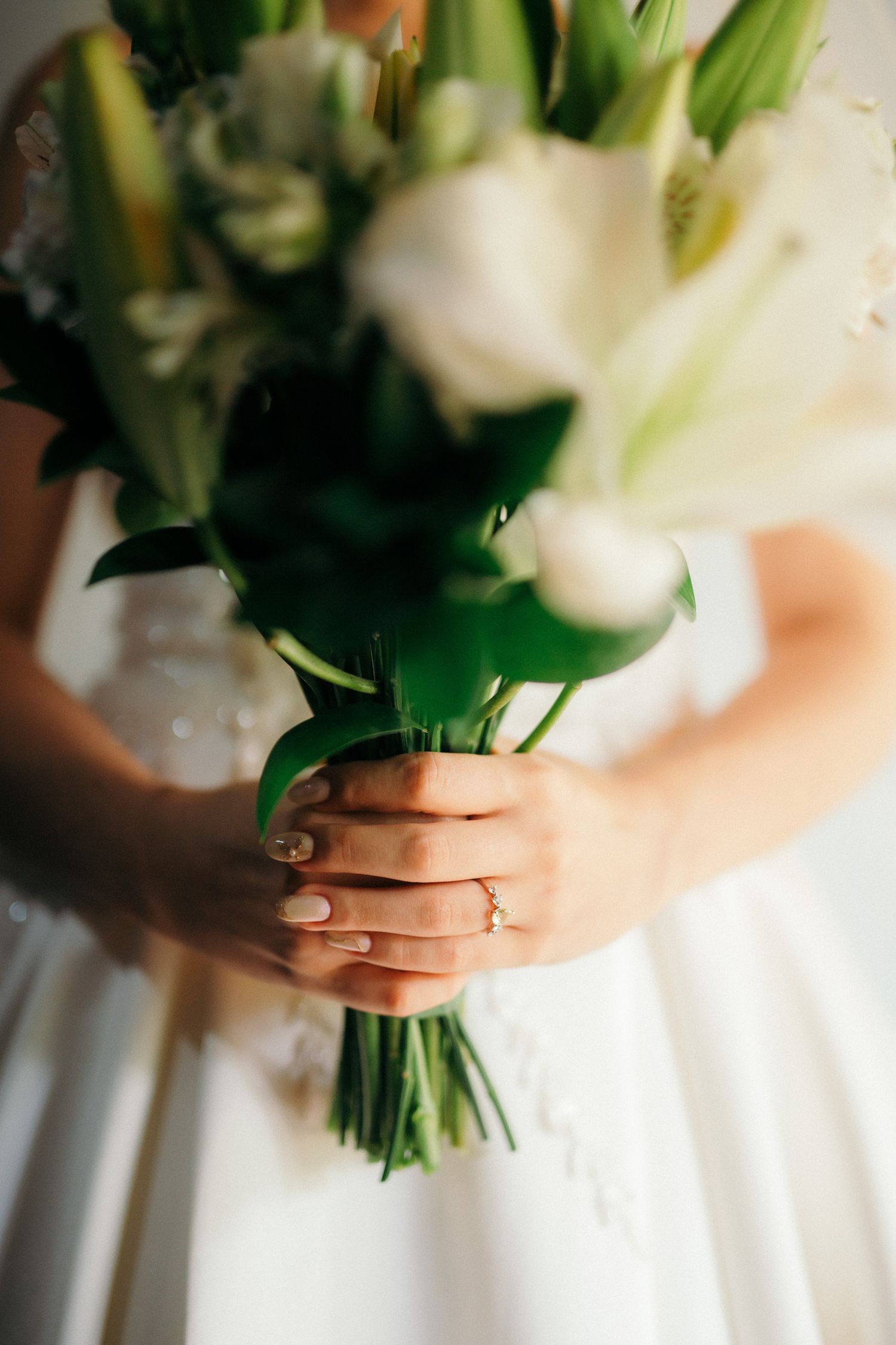 Close-up of bride holding white lily and tulip wedding bouquet with manicured hands.