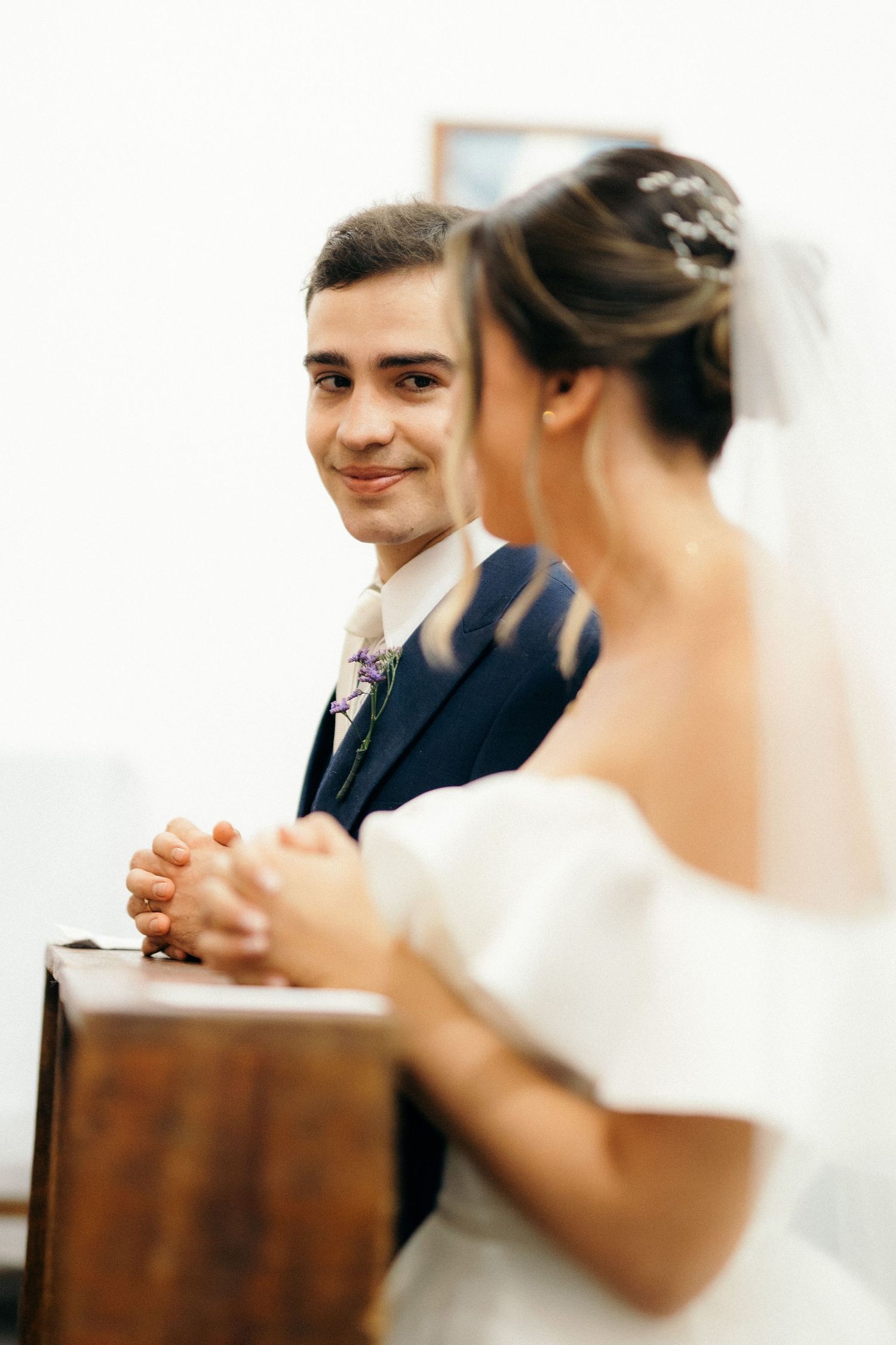 Smiling groom in navy suit gazes at bride in white off-shoulder dress during a church wedding ceremony.