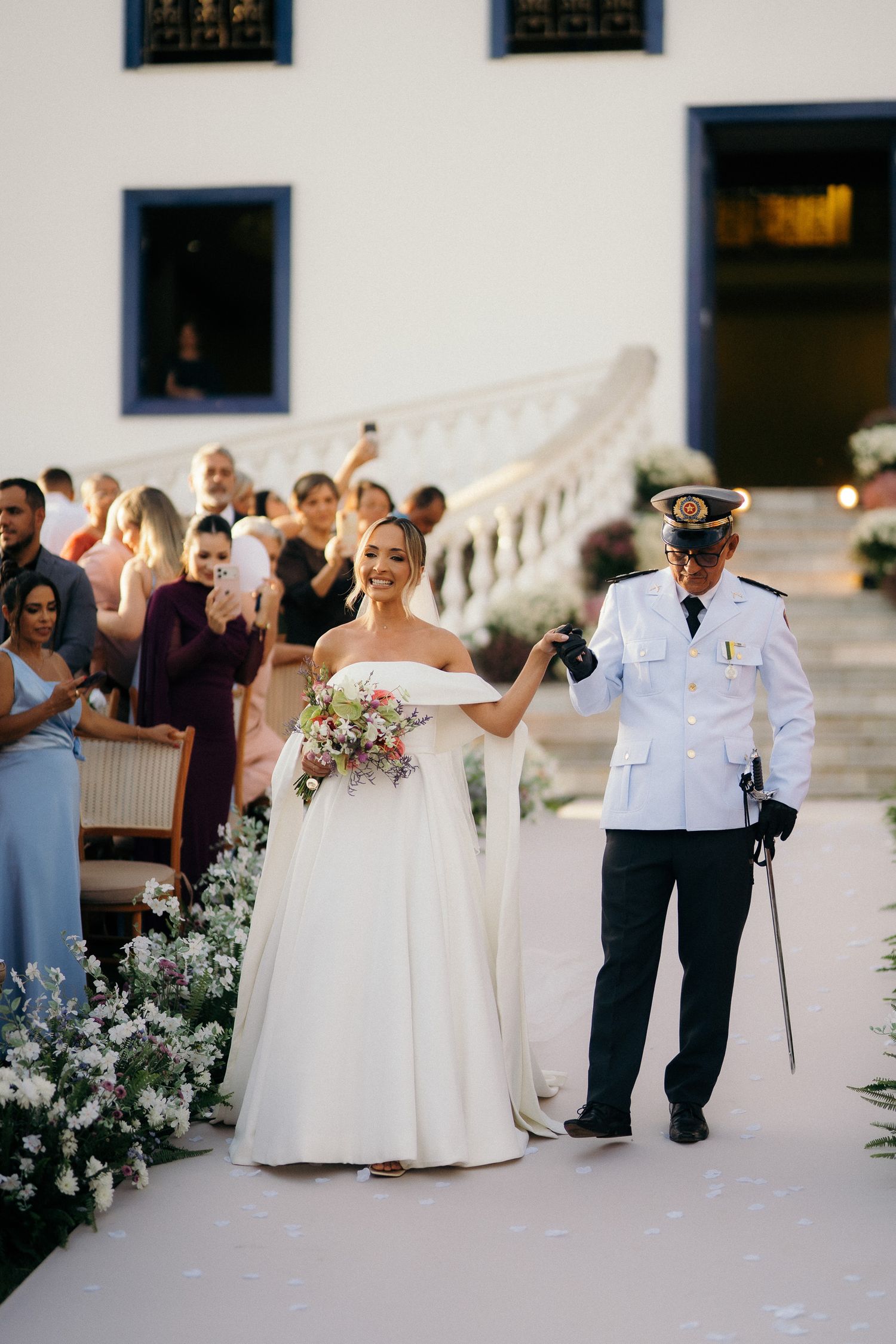 Smiling bride in off-shoulder white gown walks aisle with military officer escort holding colorful bouquet.