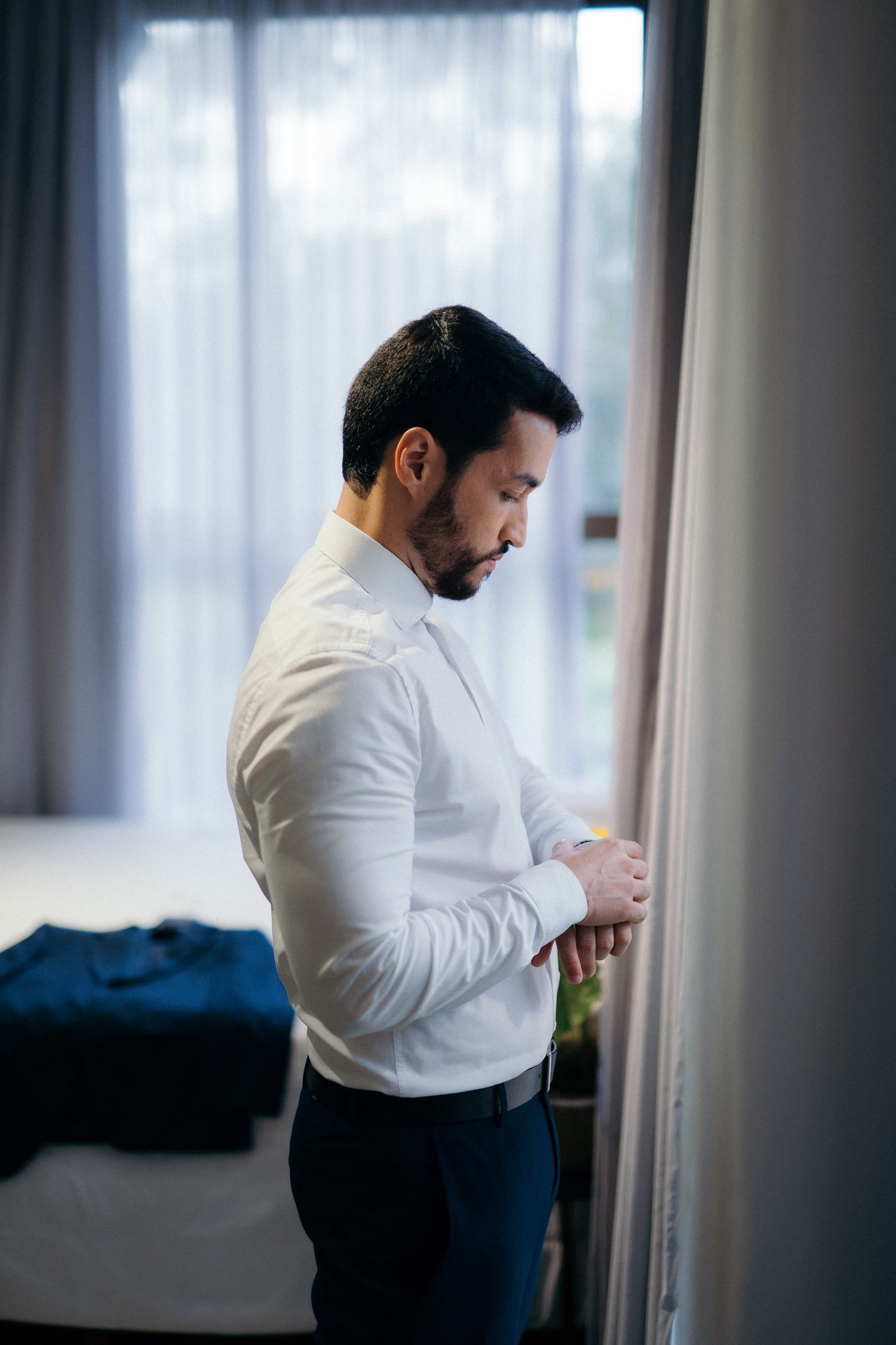 Man in white dress shirt buttoning cuffs near window, getting dressed in modern bedroom.