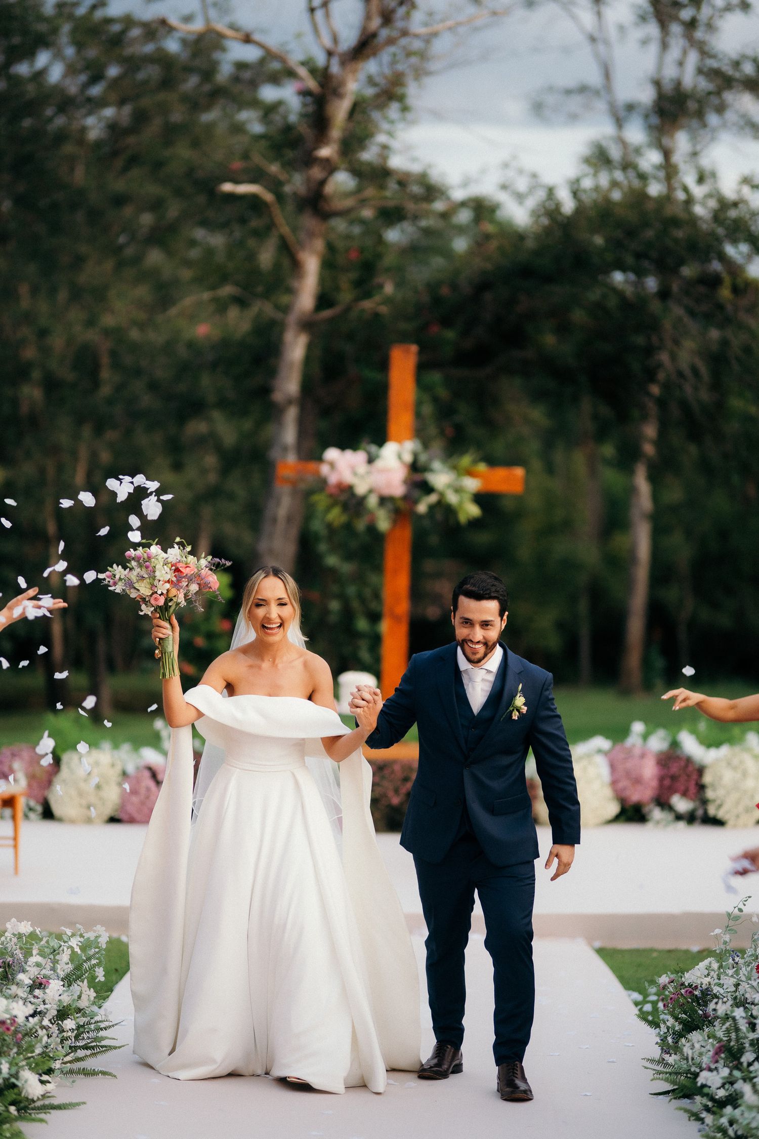 Happy newlyweds walking back down aisle after ceremony, bride raising bouquet with wooden cross and floral backdrop.
