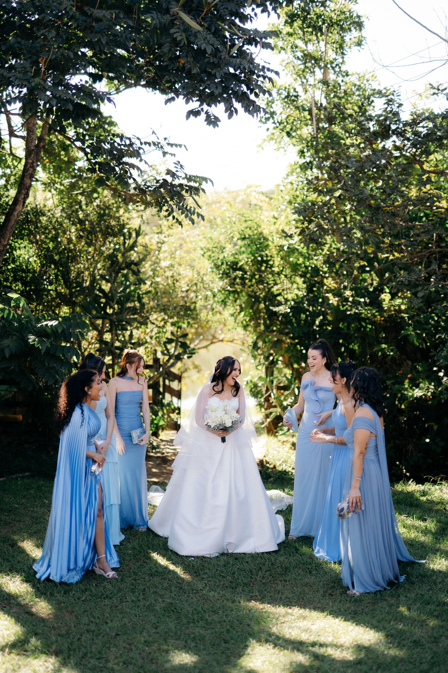 Bride in white gown surrounded by four bridesmaids in blue dresses in a lush garden.