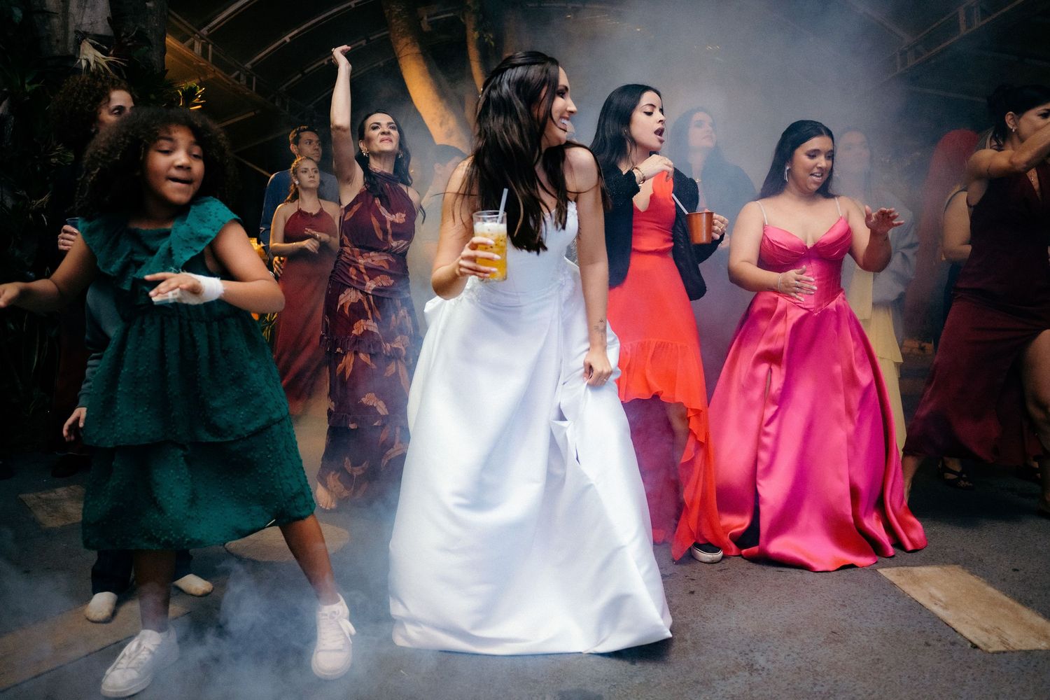 Bride in white gown dancing with colorful bridesmaids in smoke-filled wedding celebration.