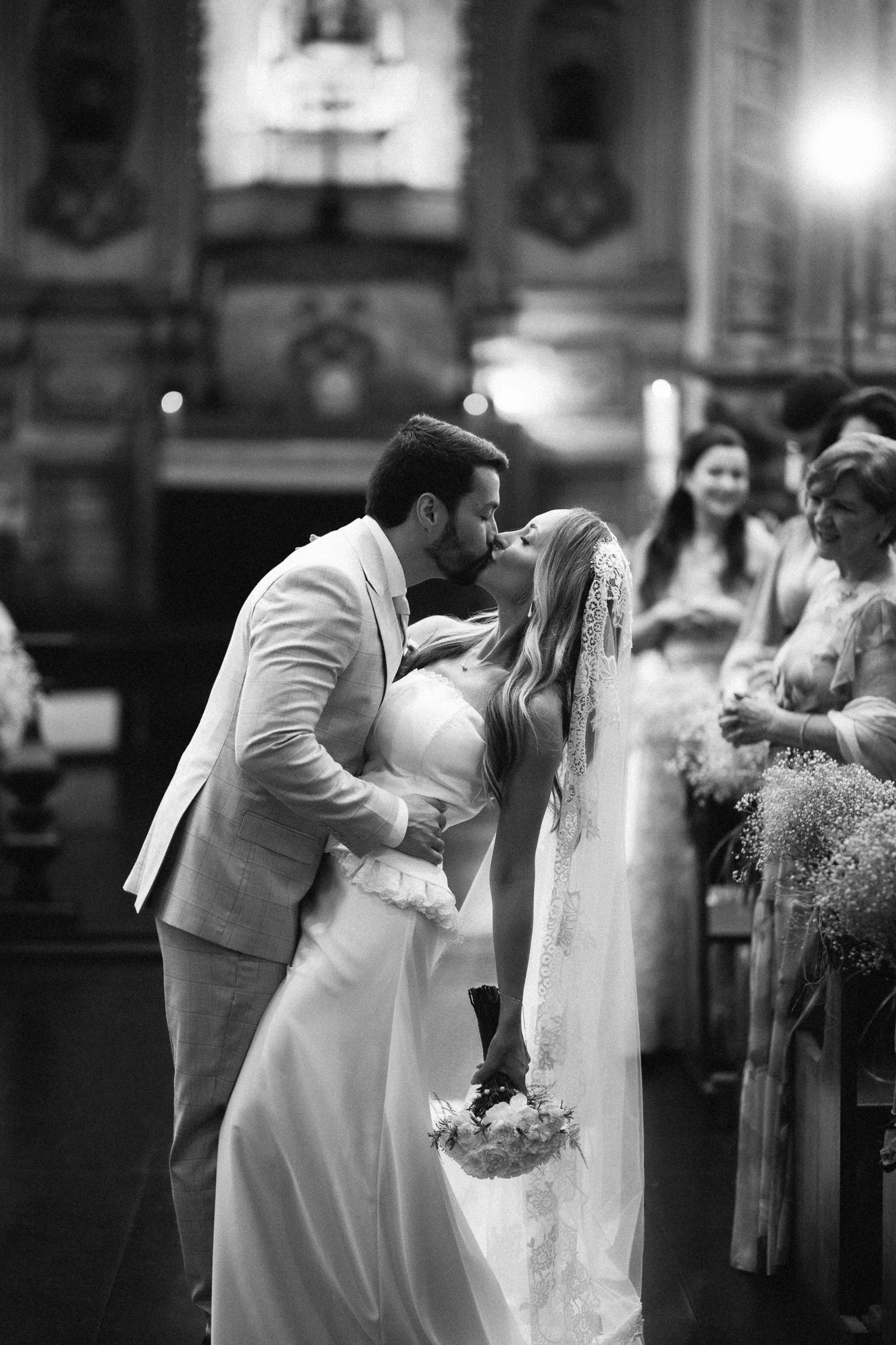 Black and white photo of bride and groom sharing first kiss at church ceremony, guests smiling in background.