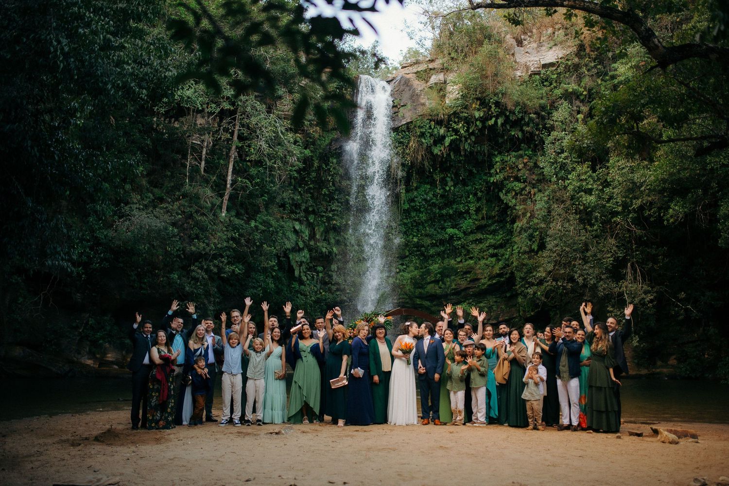 Wedding party and guests celebrate in front of a stunning waterfall in a lush tropical forest setting.