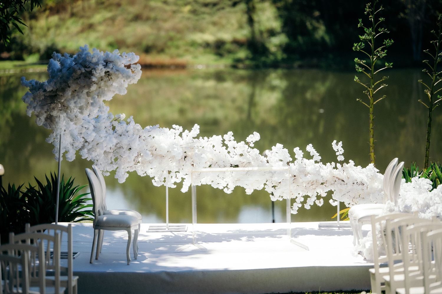 Elegant outdoor wedding ceremony setup with white floral arch and chairs beside a serene pond.