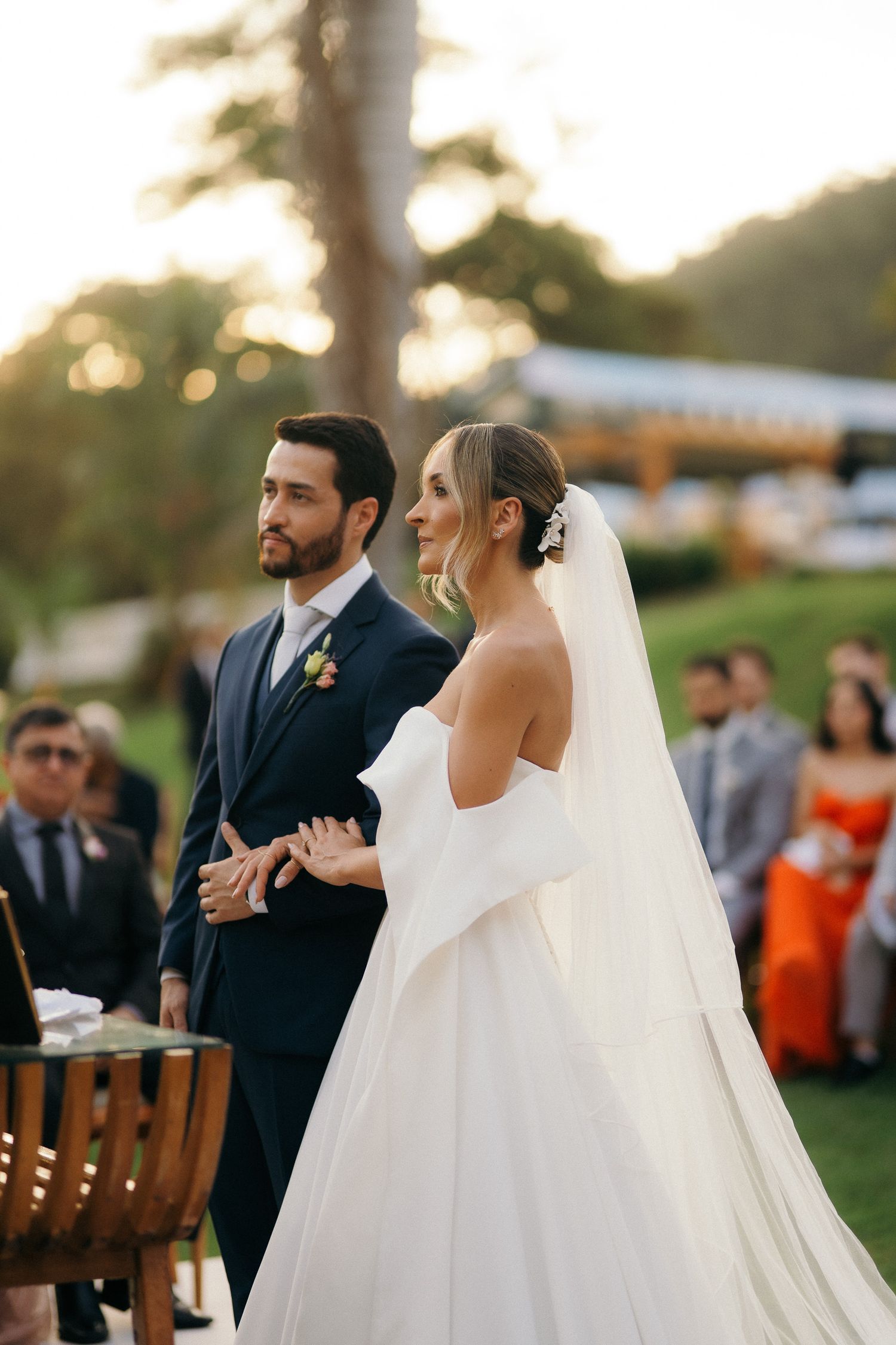 Bride in off-shoulder white gown and groom in navy suit stand together during outdoor wedding ceremony.