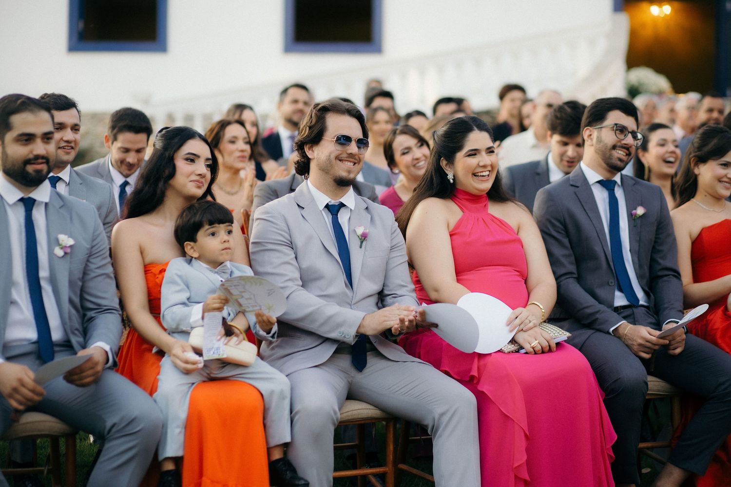 Wedding guests smiling and laughing during outdoor daytime ceremony seated in rows.