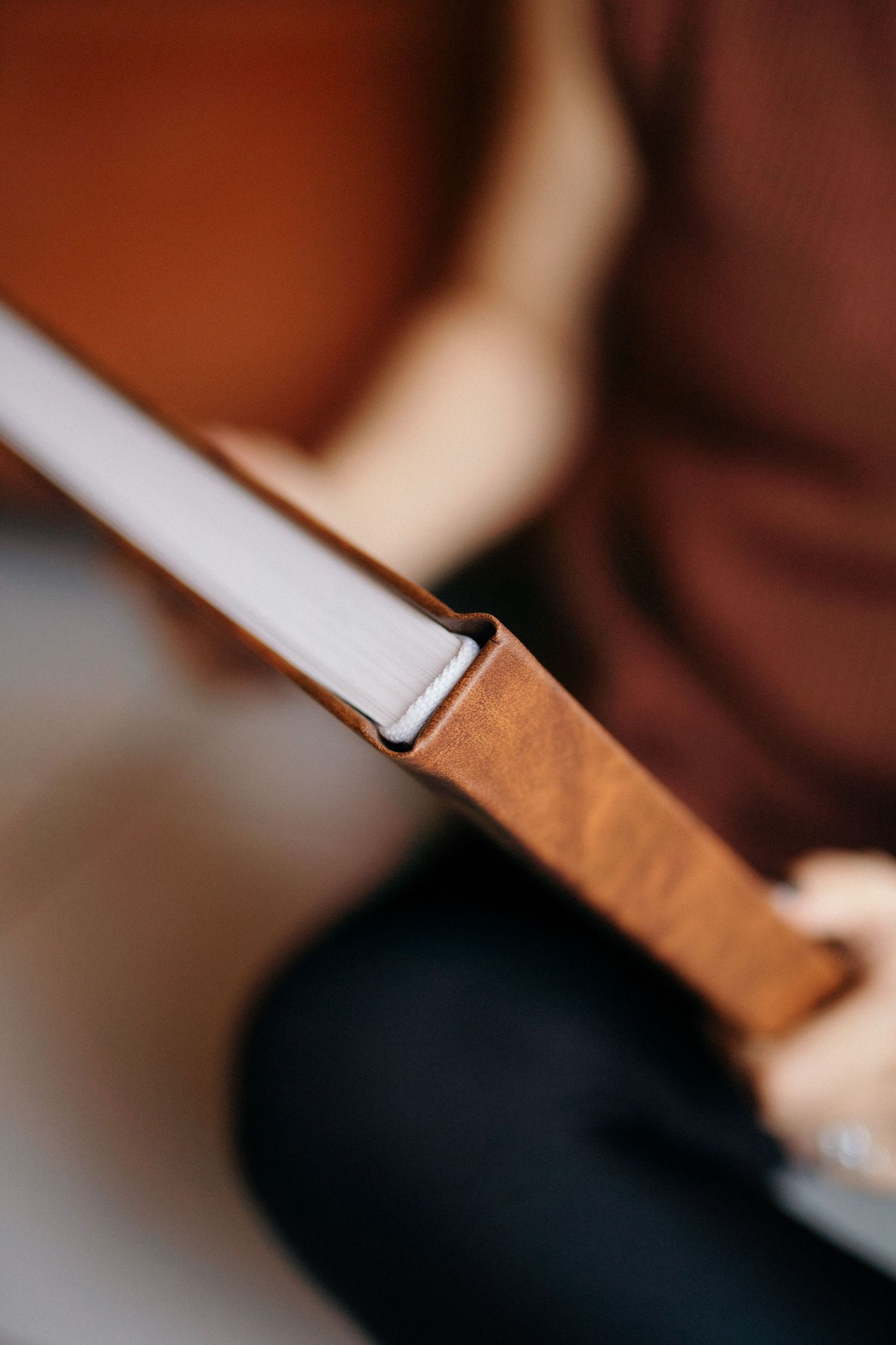 Close-up of a tan leather-bound book spine held by a person wearing a brown sweater and dark jeans.