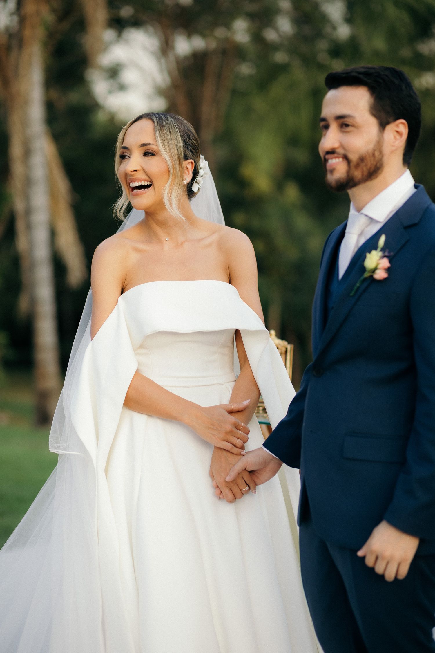 Laughing bride in strapless white gown holds hands with groom in navy suit outdoors.