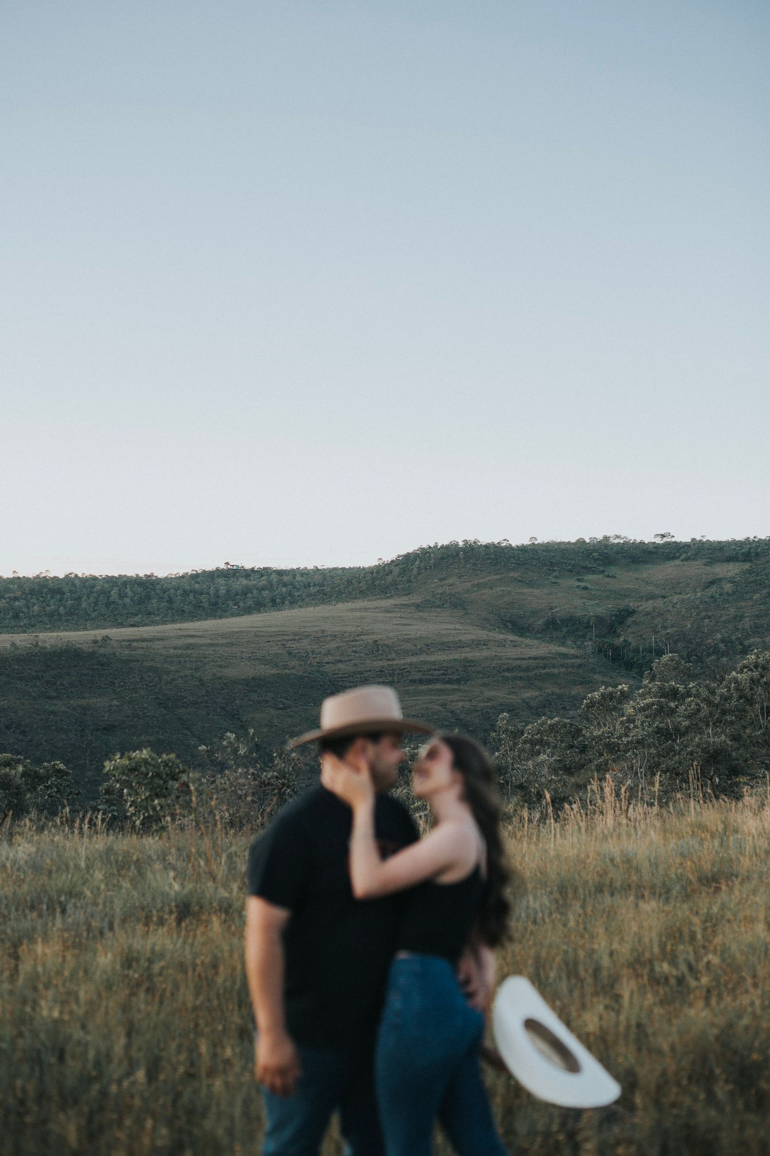 A couple shares a tender kiss in a golden field, the man wearing a tan cowboy hat against a scenic hillside.