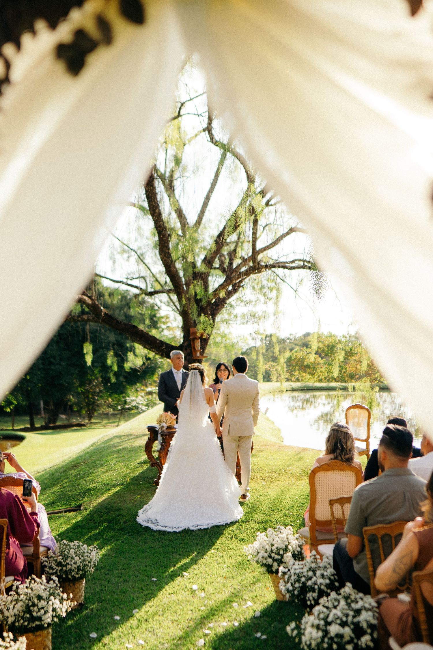 Outdoor wedding ceremony on green lawn with bride and groom at altar, framed by white draping, large tree, and pond backdrop.