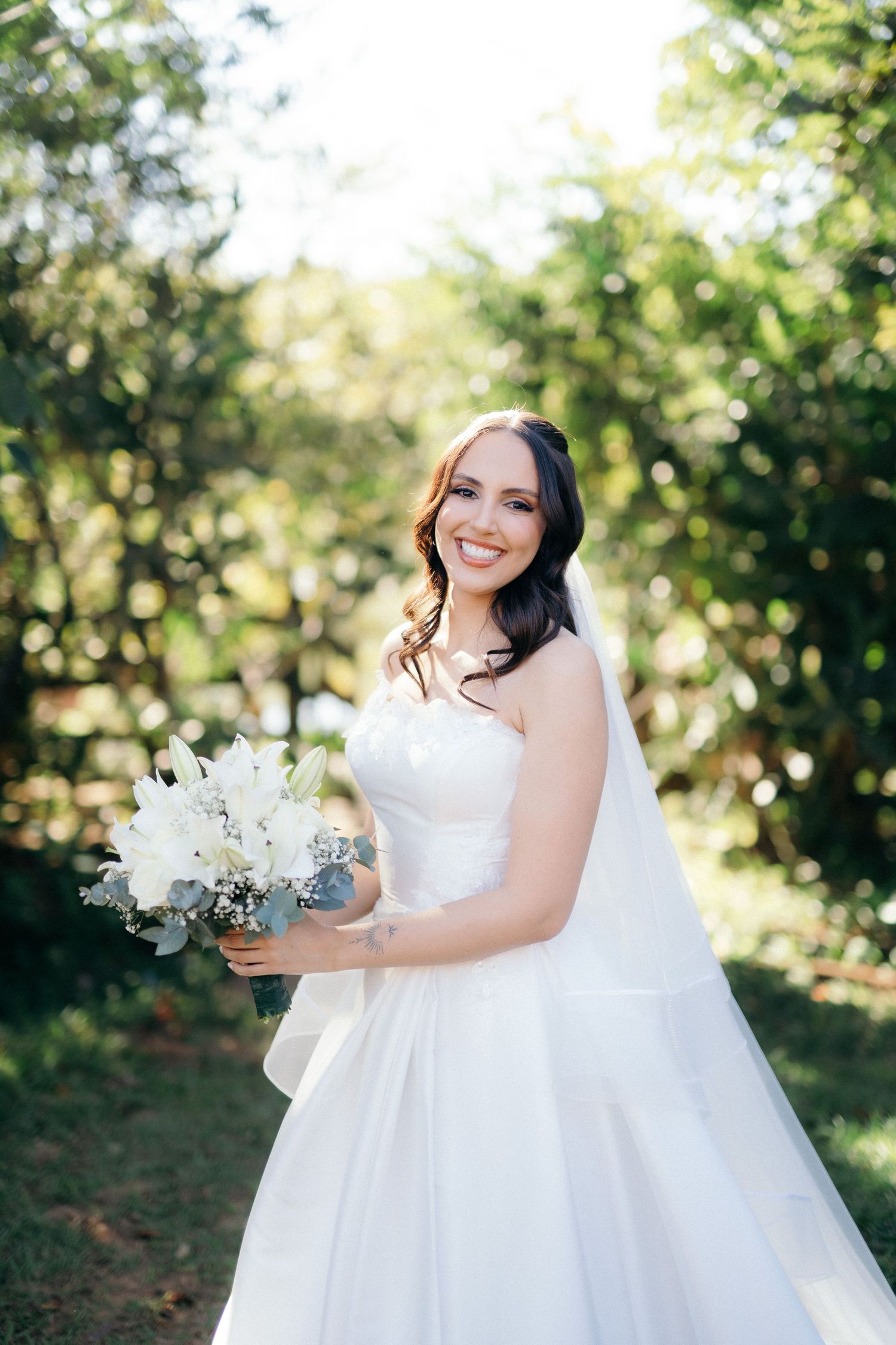 Smiling bride in white strapless gown holding white floral bouquet outdoors in green garden setting.