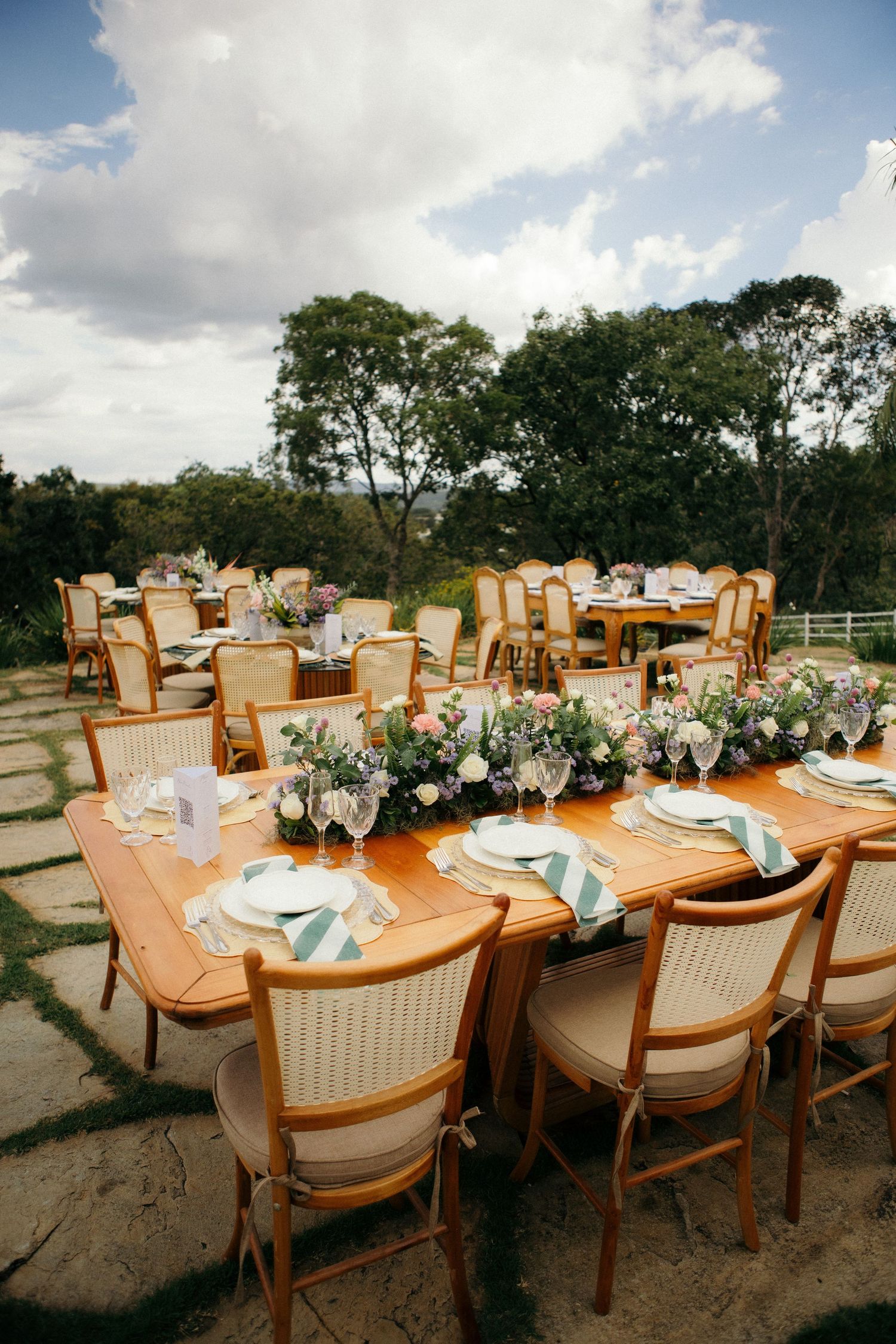 Elegant outdoor wedding reception with wooden tables, floral centerpieces, gold chairs, and scenic garden backdrop.