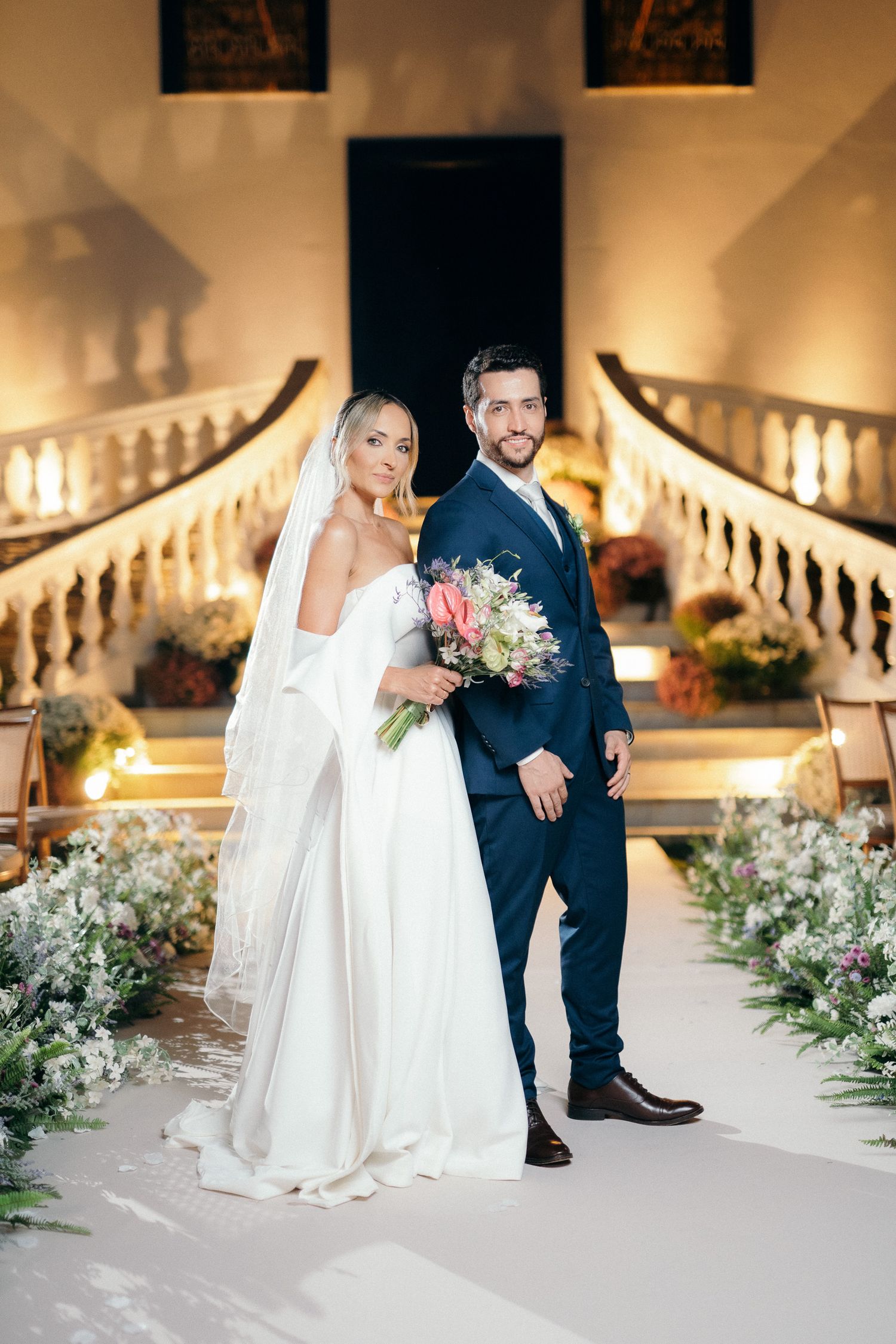 Elegant bride and groom posing on white aisle with grand staircase and warm floral lighting at night.