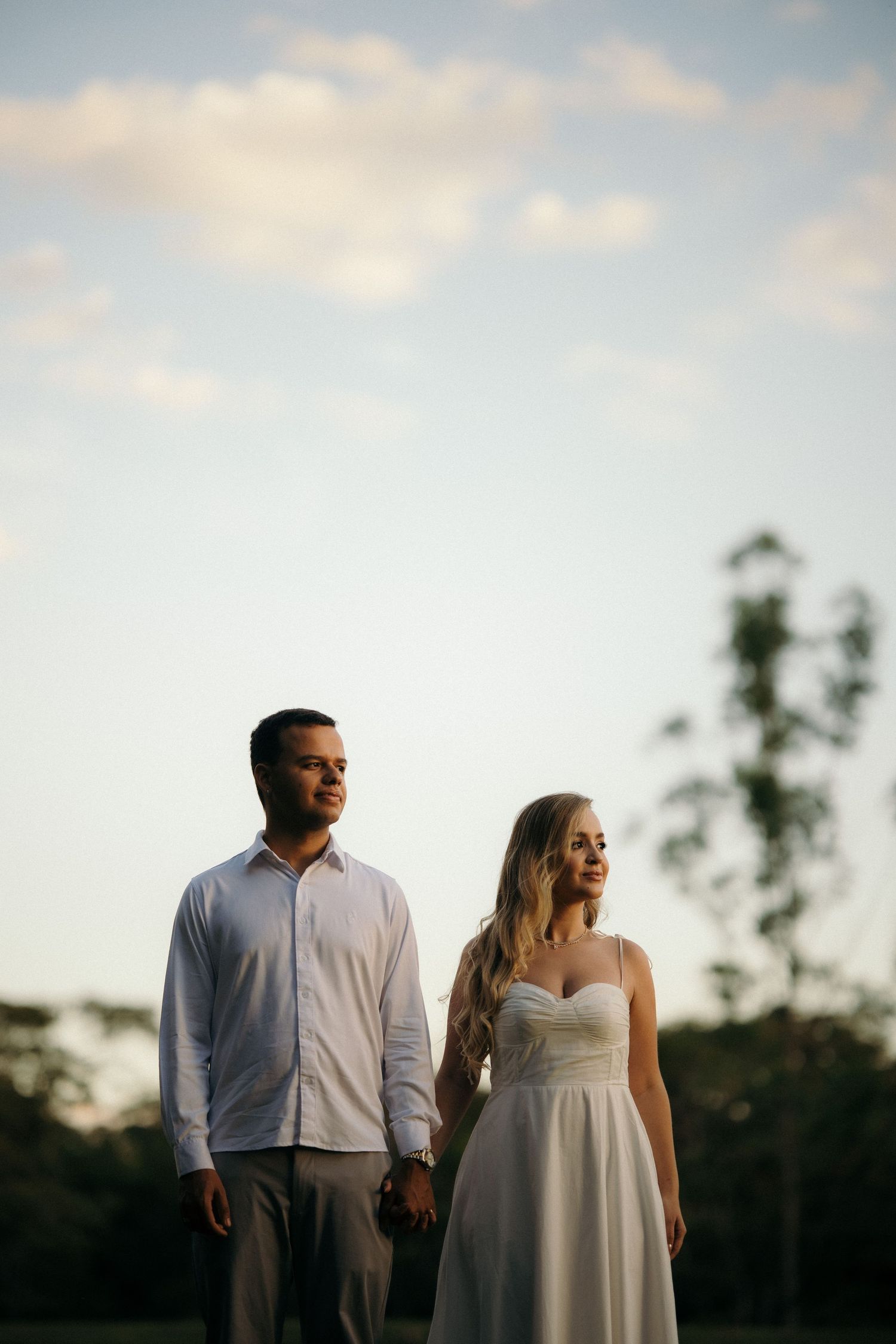 Couple in elegant attire holding hands outdoors at sunset with a cloudy sky and trees in the background.