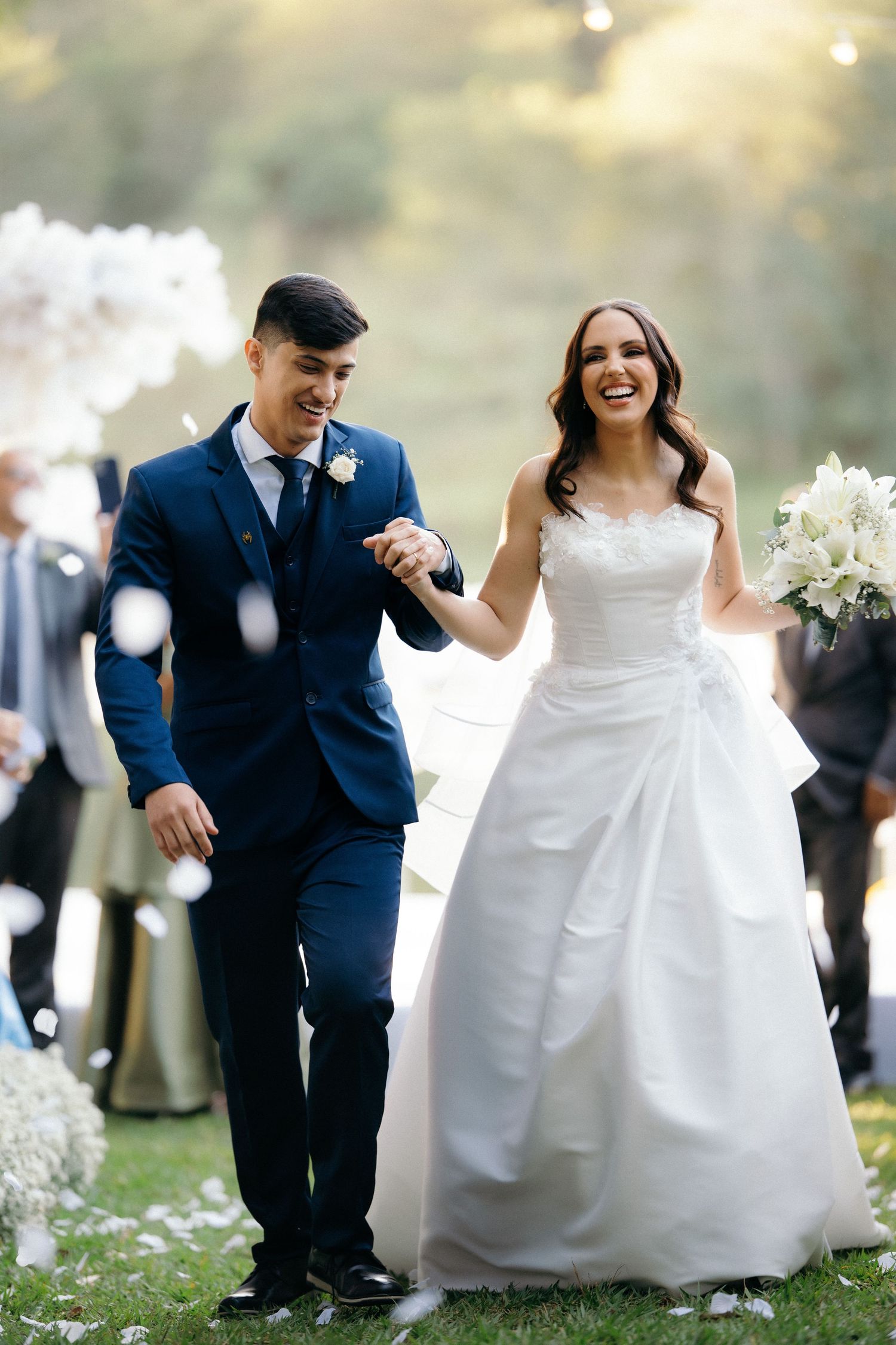 Laughing newlyweds walk through confetti shower outdoors after wedding ceremony.