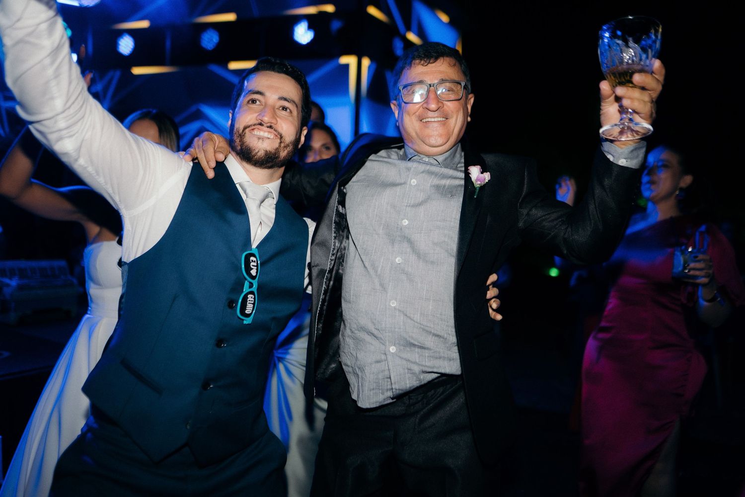 Two men celebrate joyfully raising glasses at a wedding reception under blue lights.