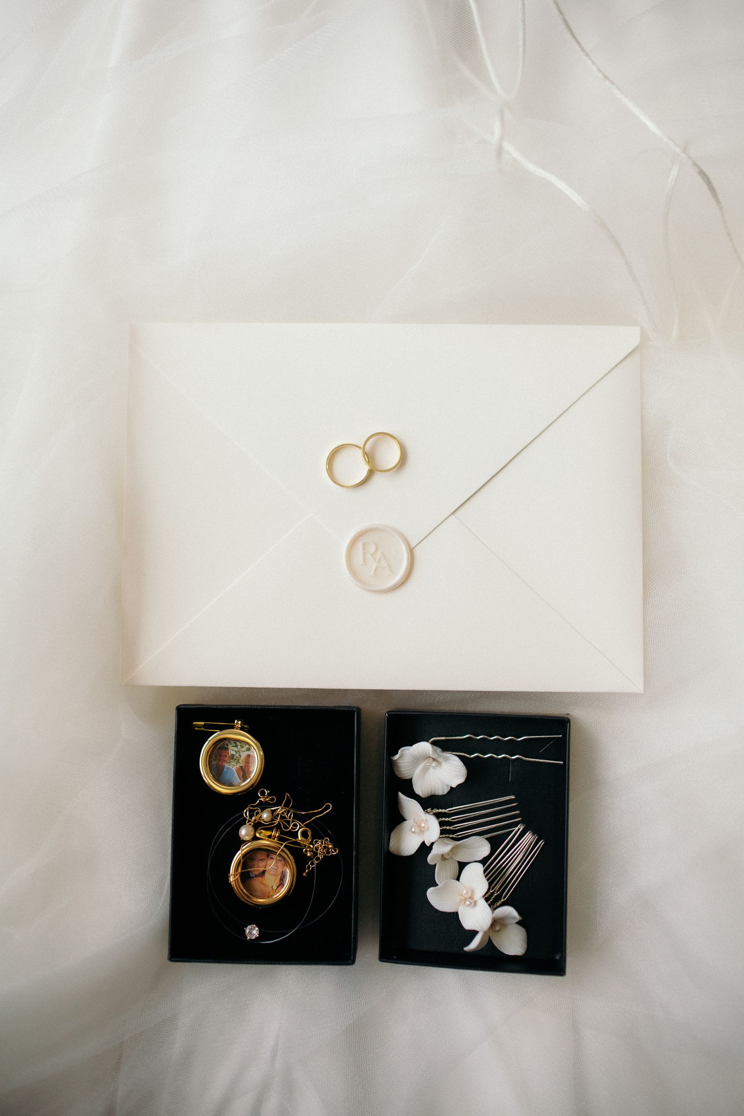 Elegant wedding flat lay with gold rings on ivory envelope, wax seal, locket jewelry, and white floral hair pins in black boxes.