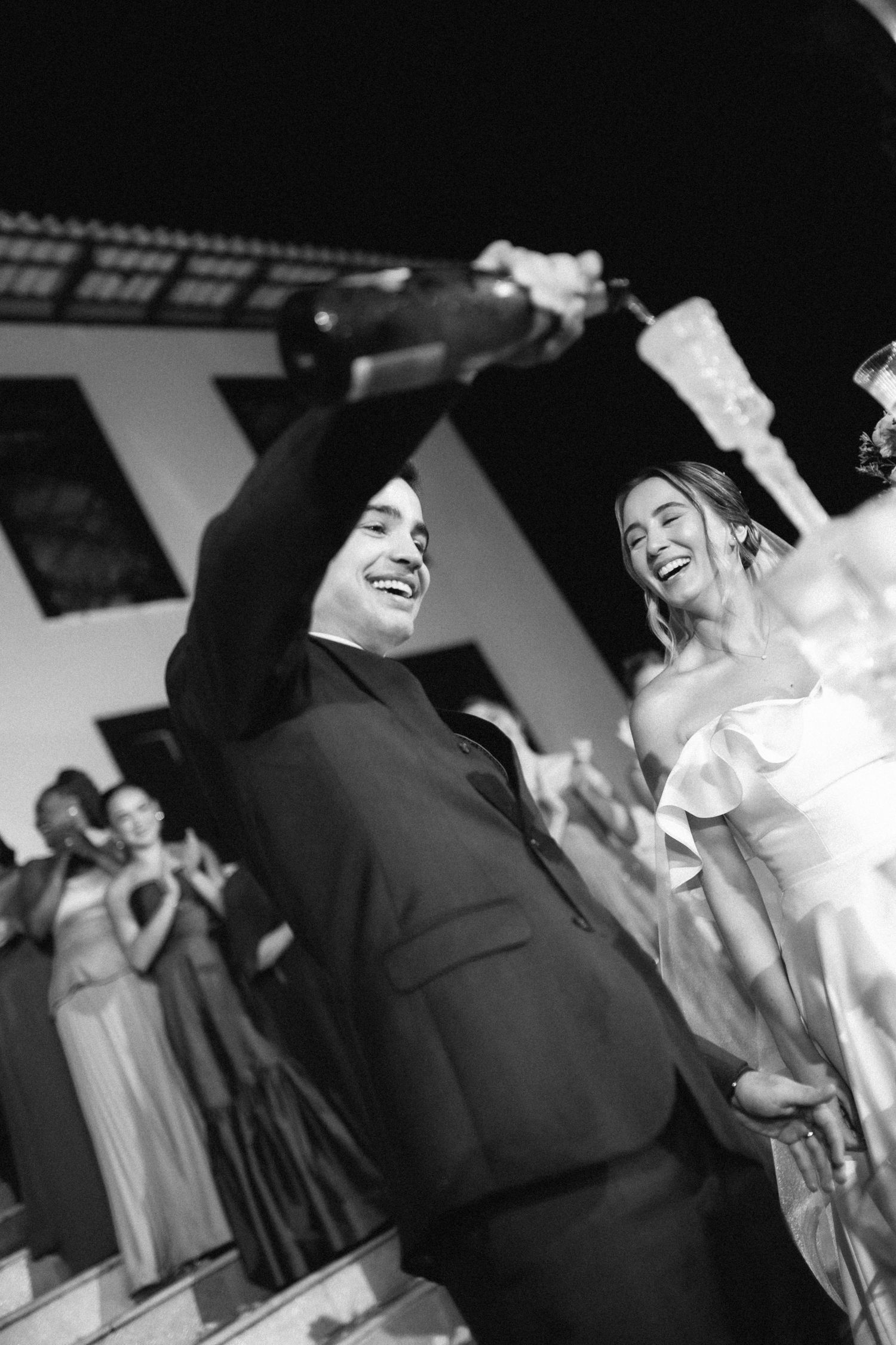 Groom laughing while popping champagne bottle at wedding reception as bride smiles behind him.