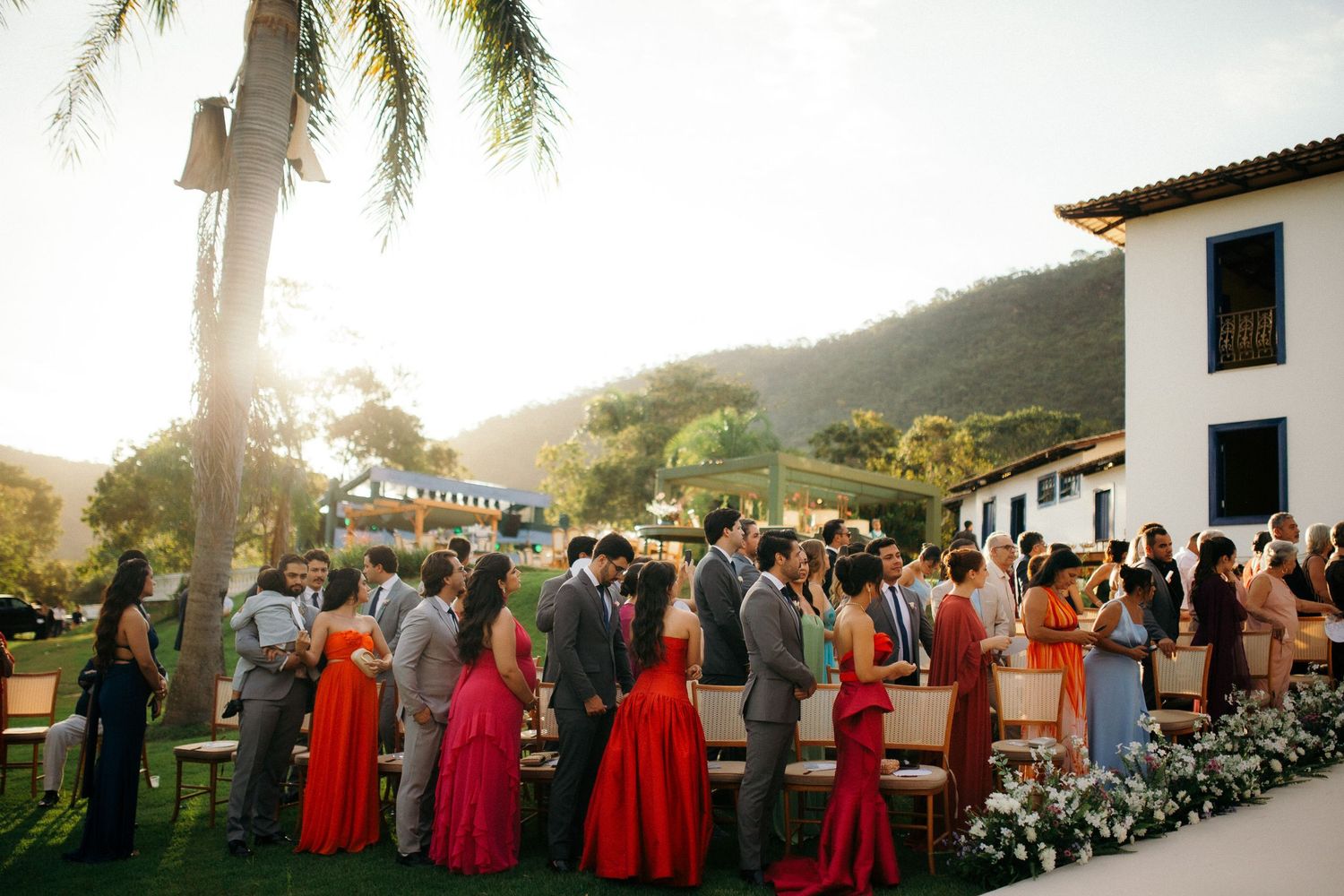 Wedding guests mingling outdoors at golden hour near a white colonial venue with palm trees.