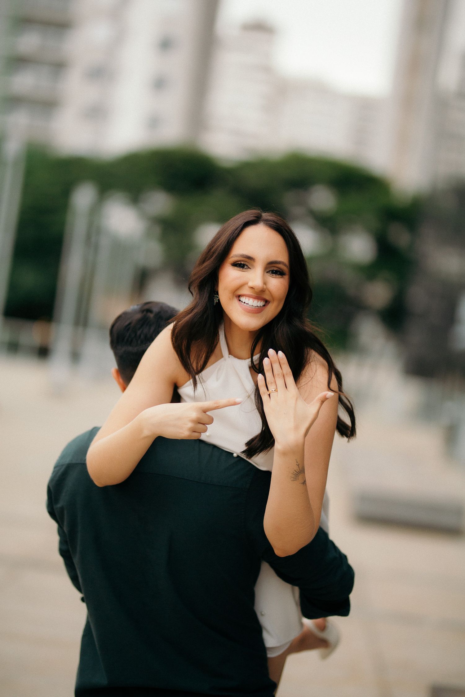 Excited woman shows off engagement ring while being lifted by partner in urban outdoor setting.