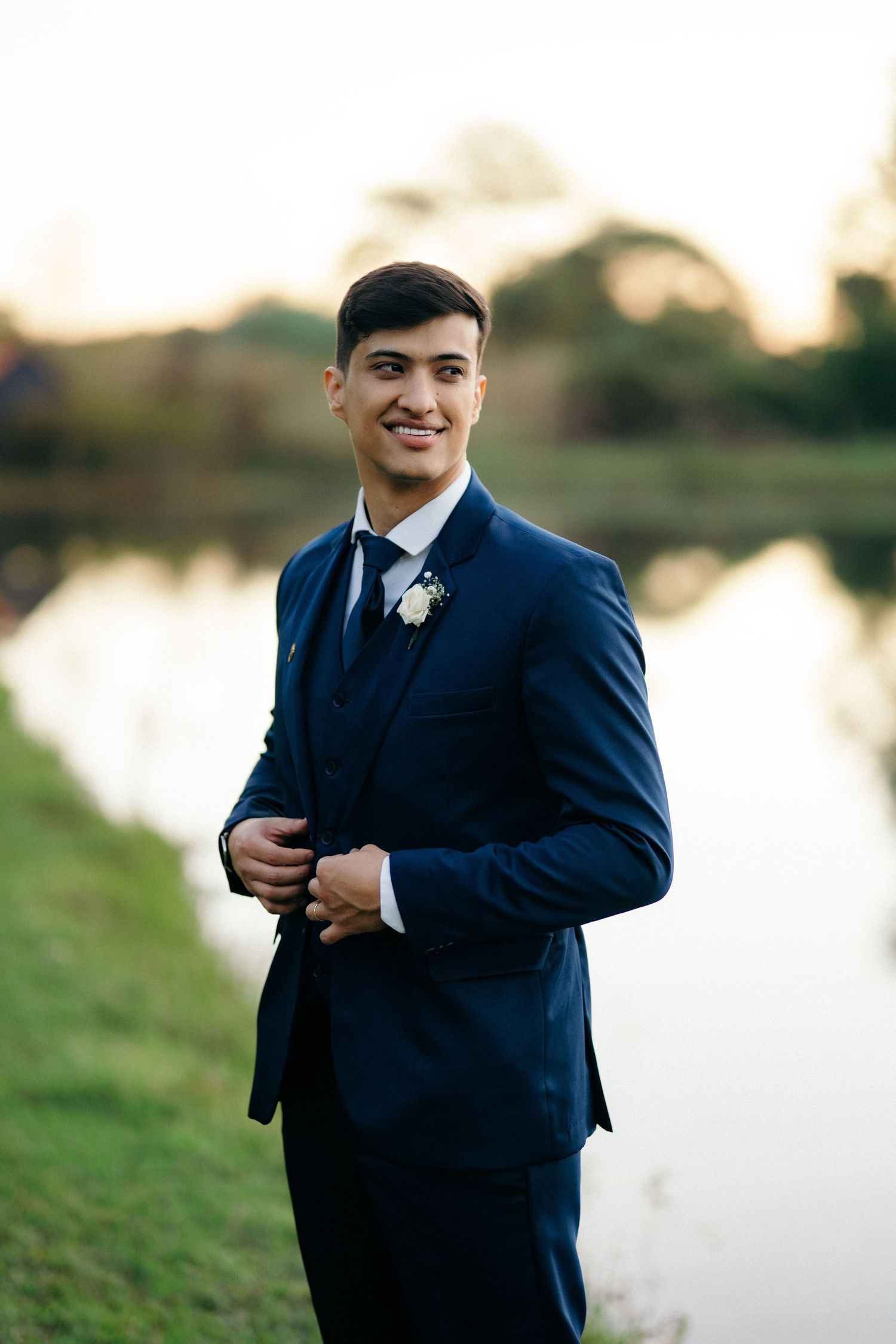 Smiling groom in a navy blue three-piece suit with boutonniere posing by a reflective pond at sunset.
