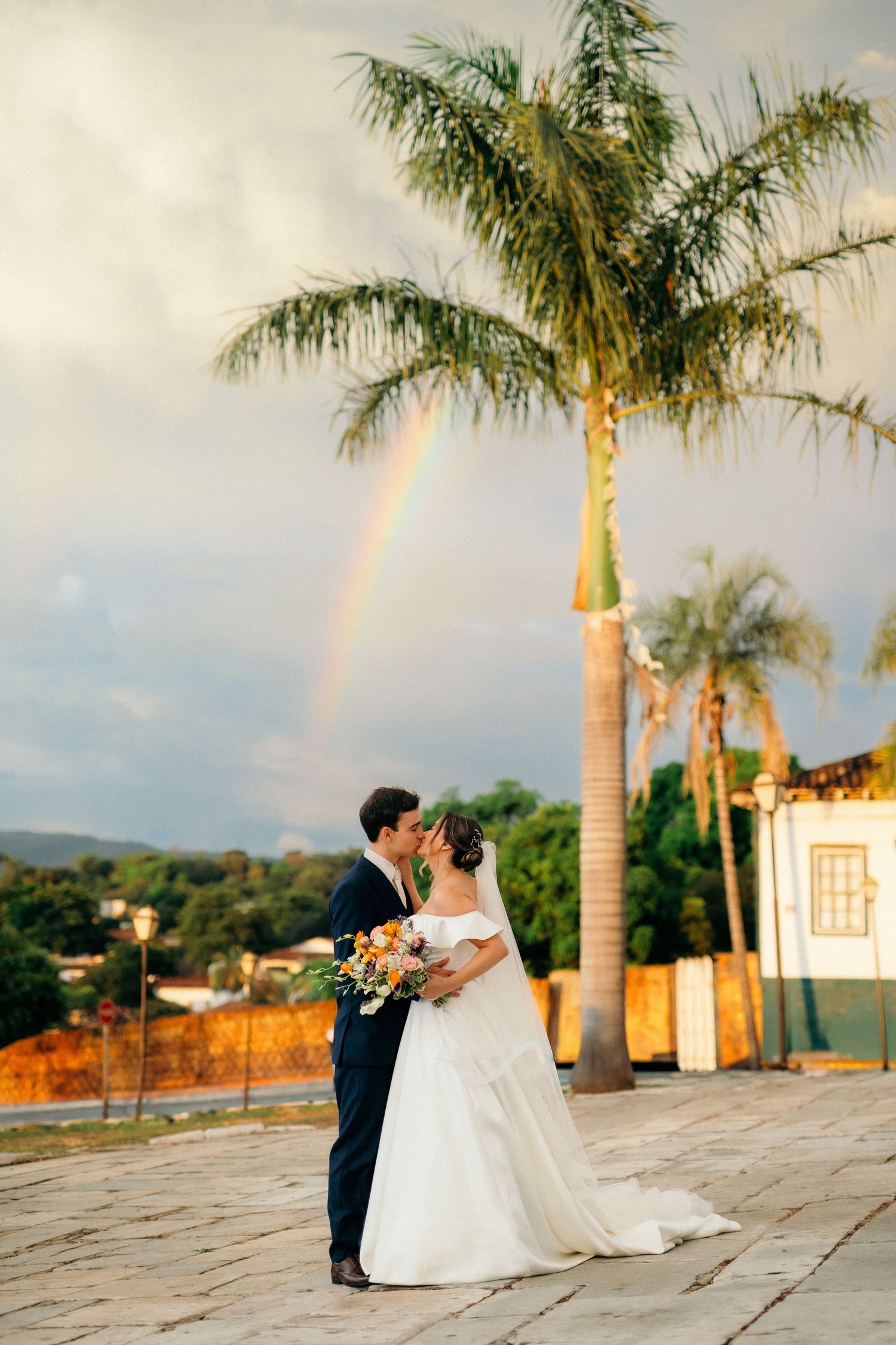 Bride and groom kissing outdoors near palm trees with a rainbow in the background.