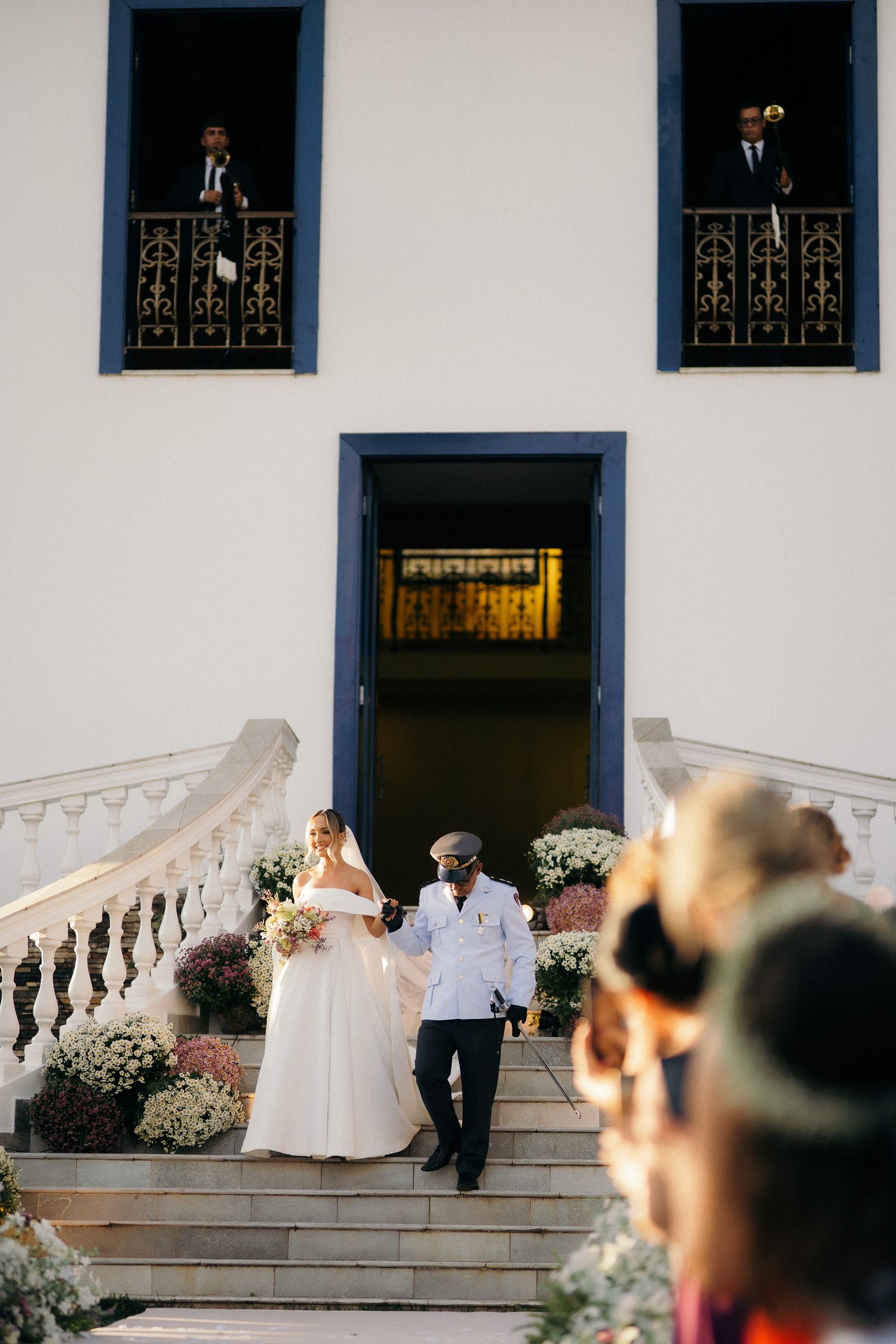 Bride descends ornate staircase with escort at elegant wedding venue while musicians play from balconies above.