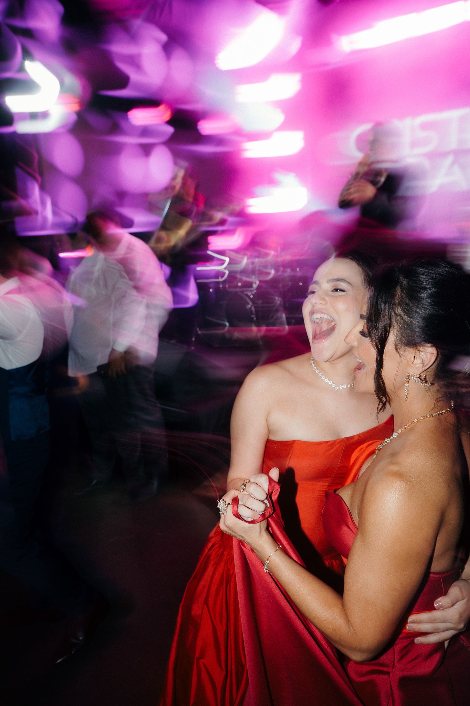 Bridesmaid in red satin gown laughs while dancing at colorful neon-lit wedding reception party.