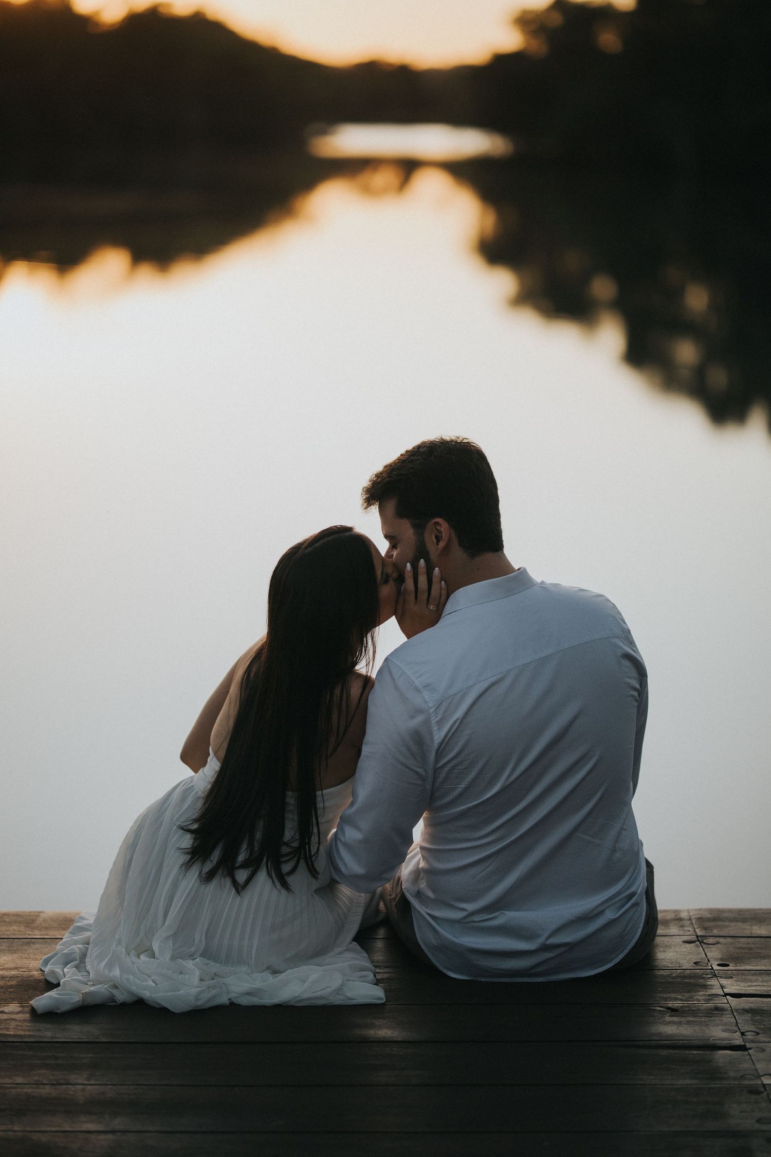 A couple shares a romantic kiss while sitting on a wooden dock overlooking a serene lake at sunset.