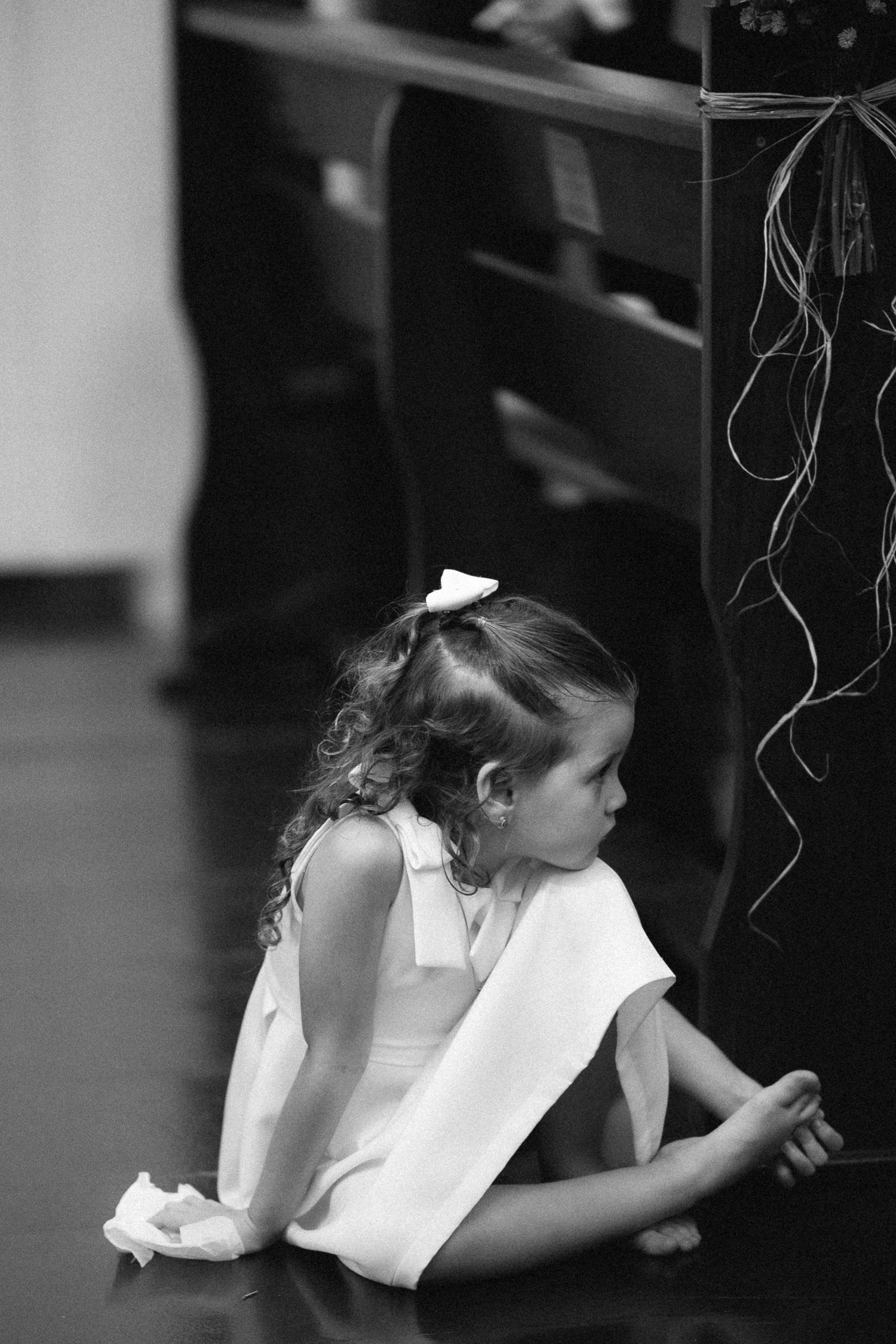 Black and white photo of a young flower girl in white dress sitting on the church floor during wedding ceremony.
