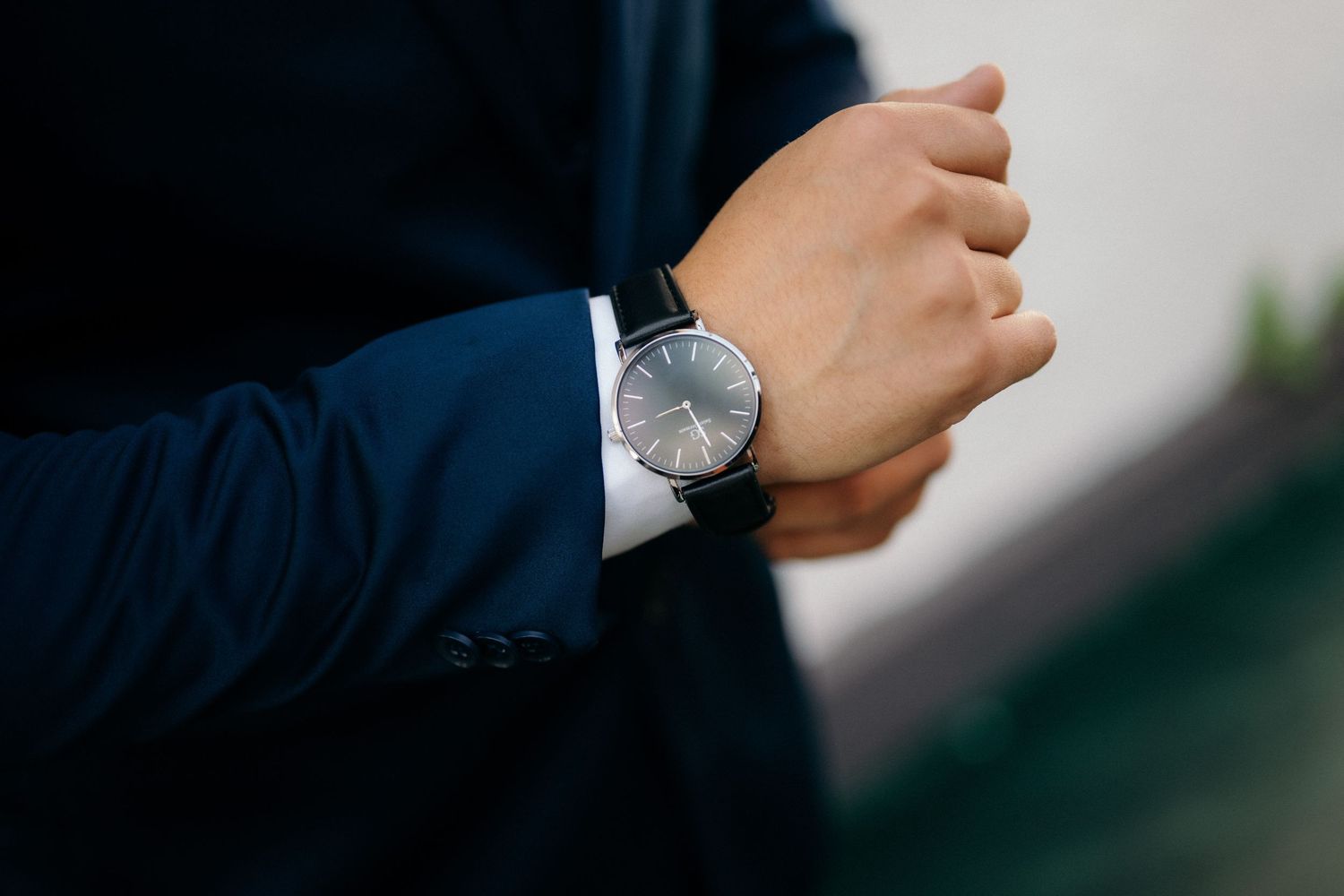 Man in navy blue suit checking time on elegant black leather strap analog wristwatch.