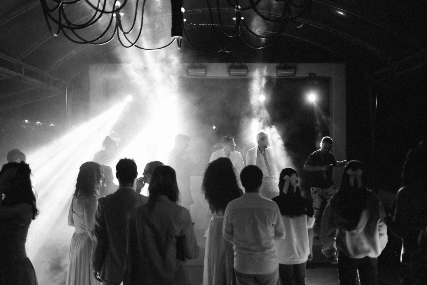 Black and white photo of wedding guests dancing under dramatic stage lights and fog at a reception.