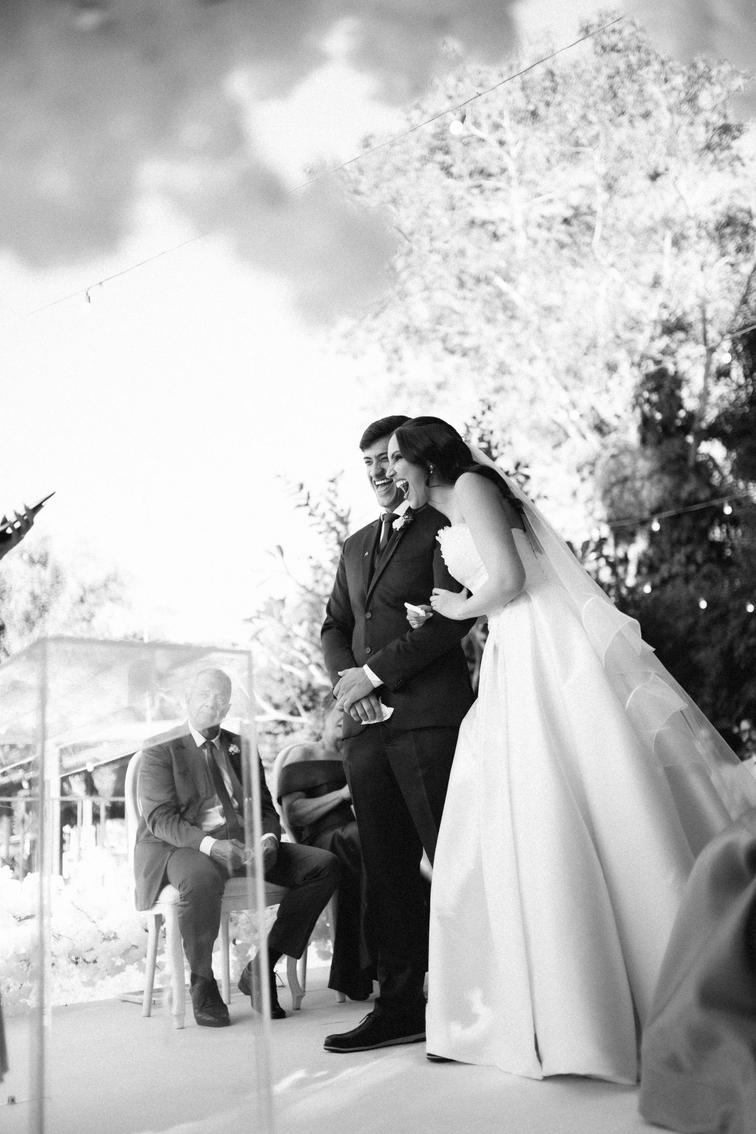 Black and white photo of a laughing bride and groom during their outdoor wedding ceremony with guests seated nearby.