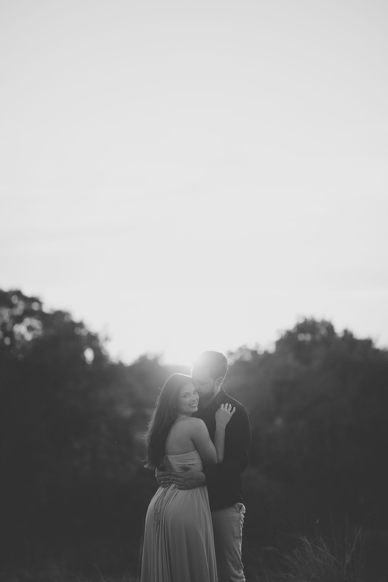 Black and white silhouette of couple embracing at sunset with glowing backlight through trees.