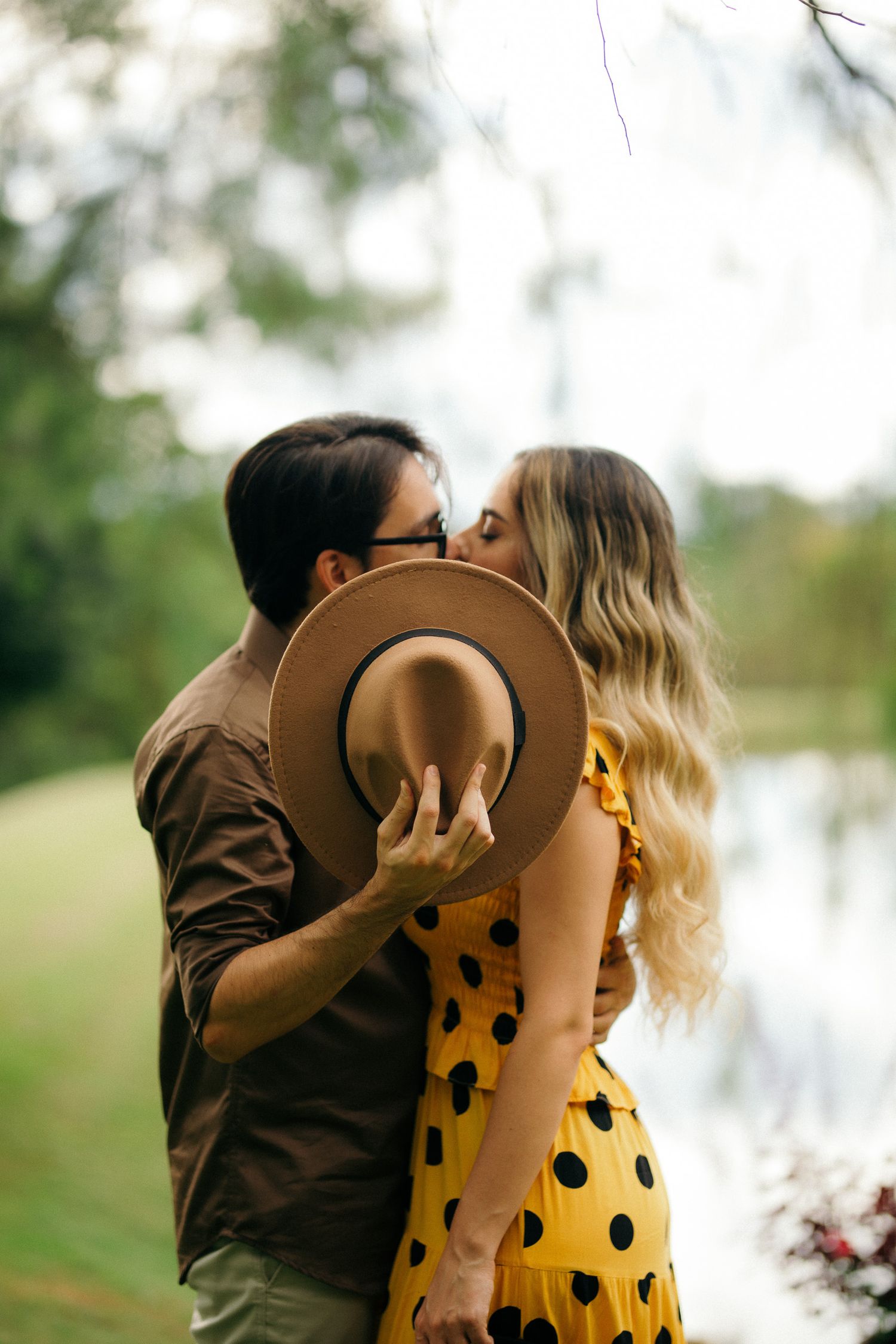 Couple sharing a romantic kiss behind a tan hat outdoors near a lake.