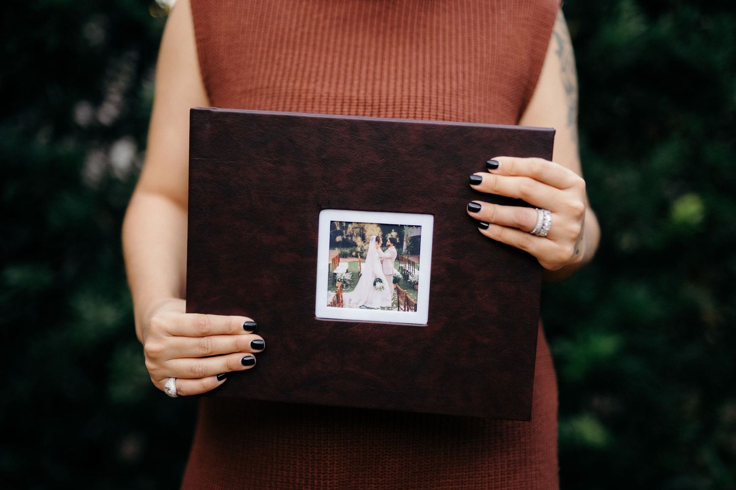 Woman in rust dress holding a dark brown leather wedding photo album with a polaroid photo on the cover.