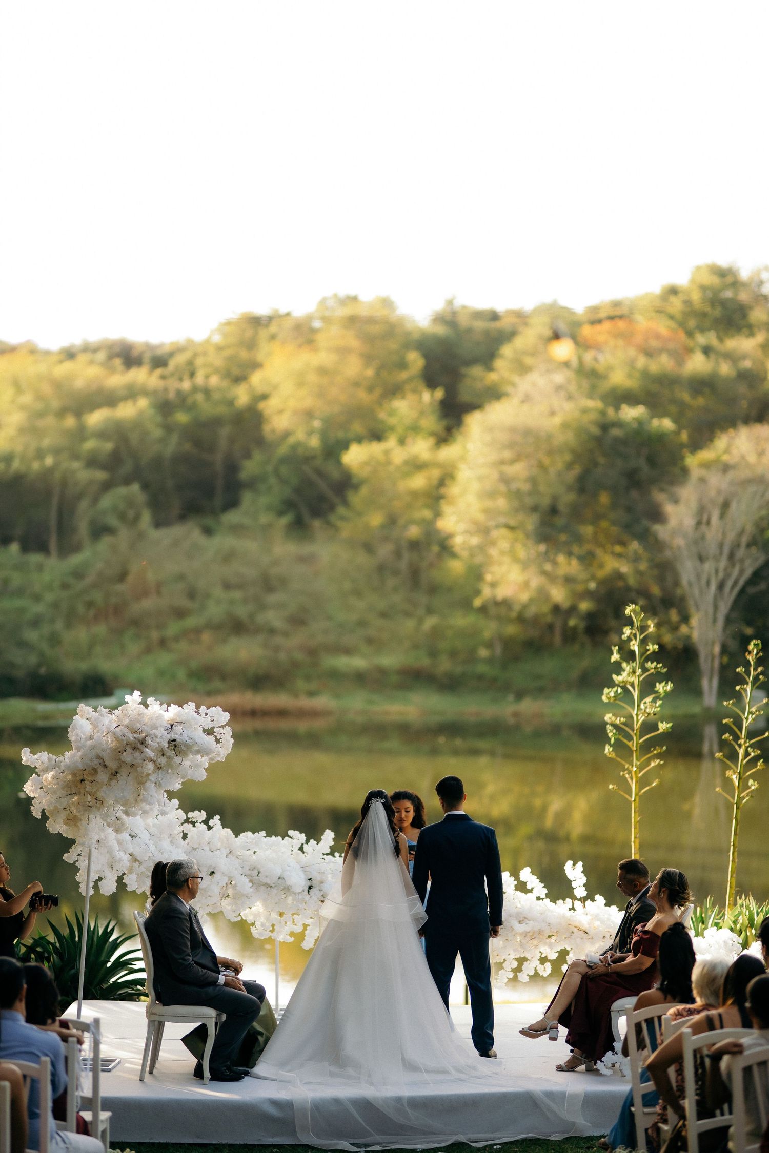 Outdoor wedding ceremony with bride and groom at altar adorned with white orchids beside a serene lake.