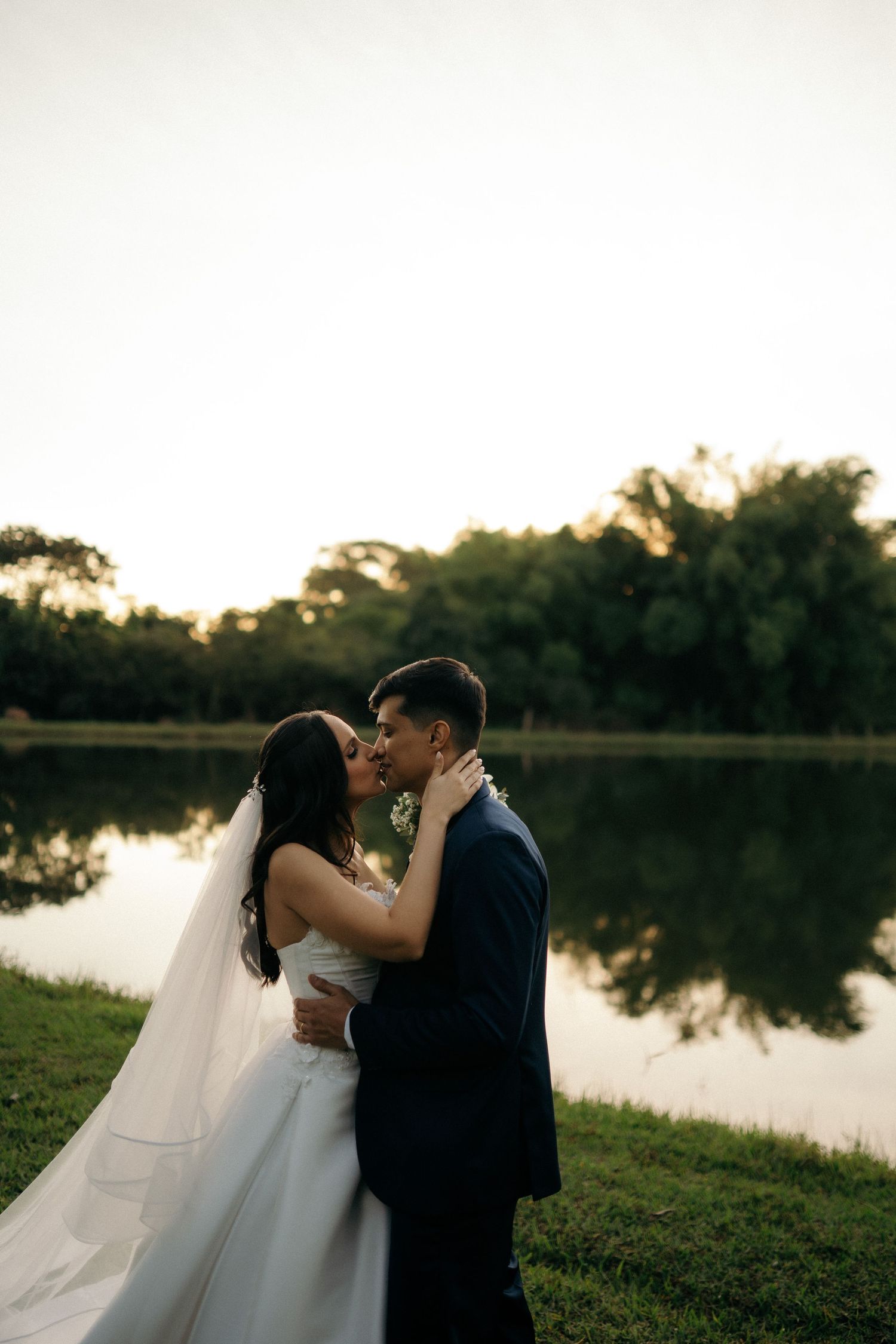 Bride and groom share romantic kiss by reflective lake at sunset during outdoor wedding.