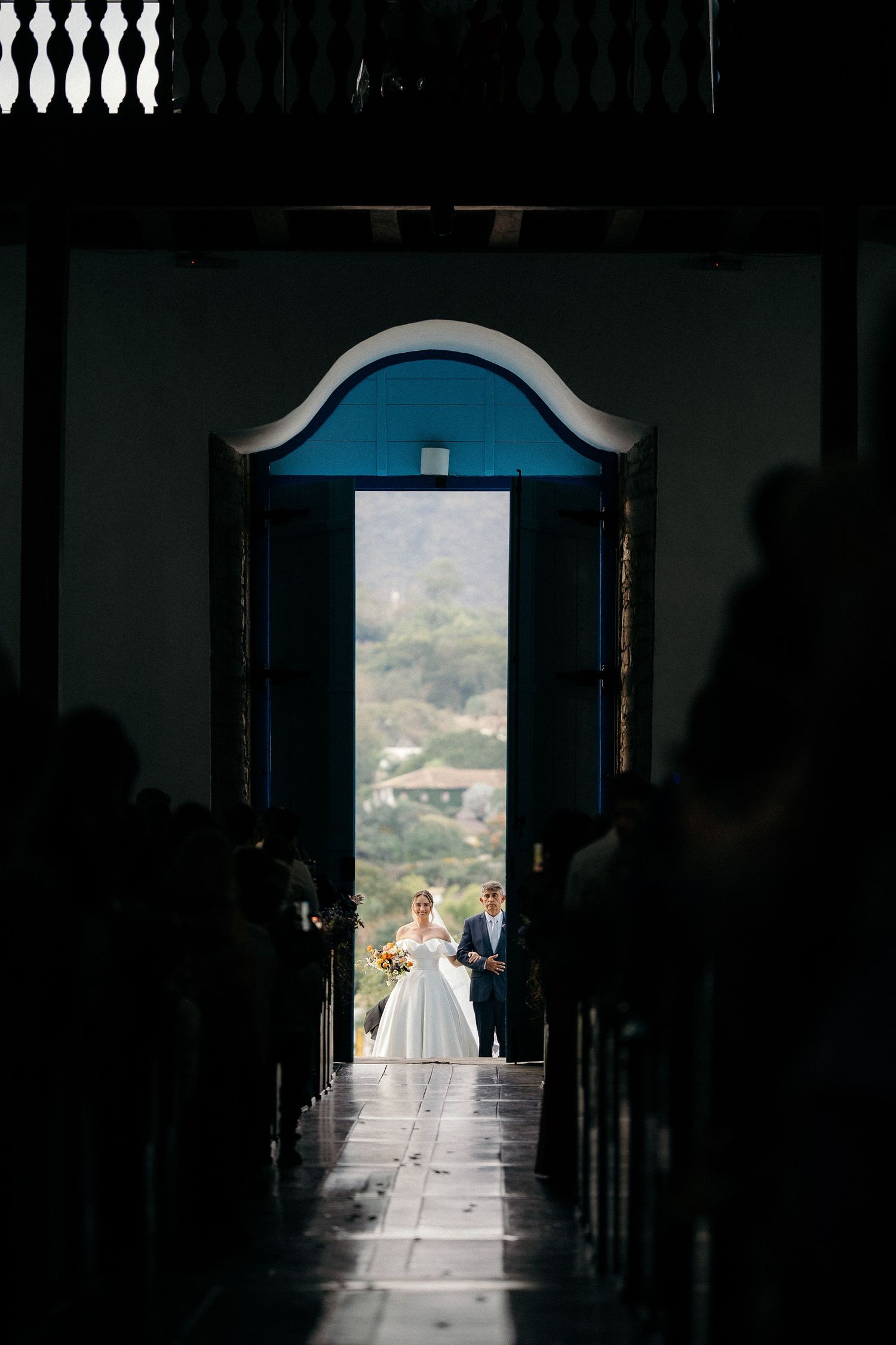 Bride and groom stand in sunlit church doorway, framed by pews and arched blue doors with mountain views.