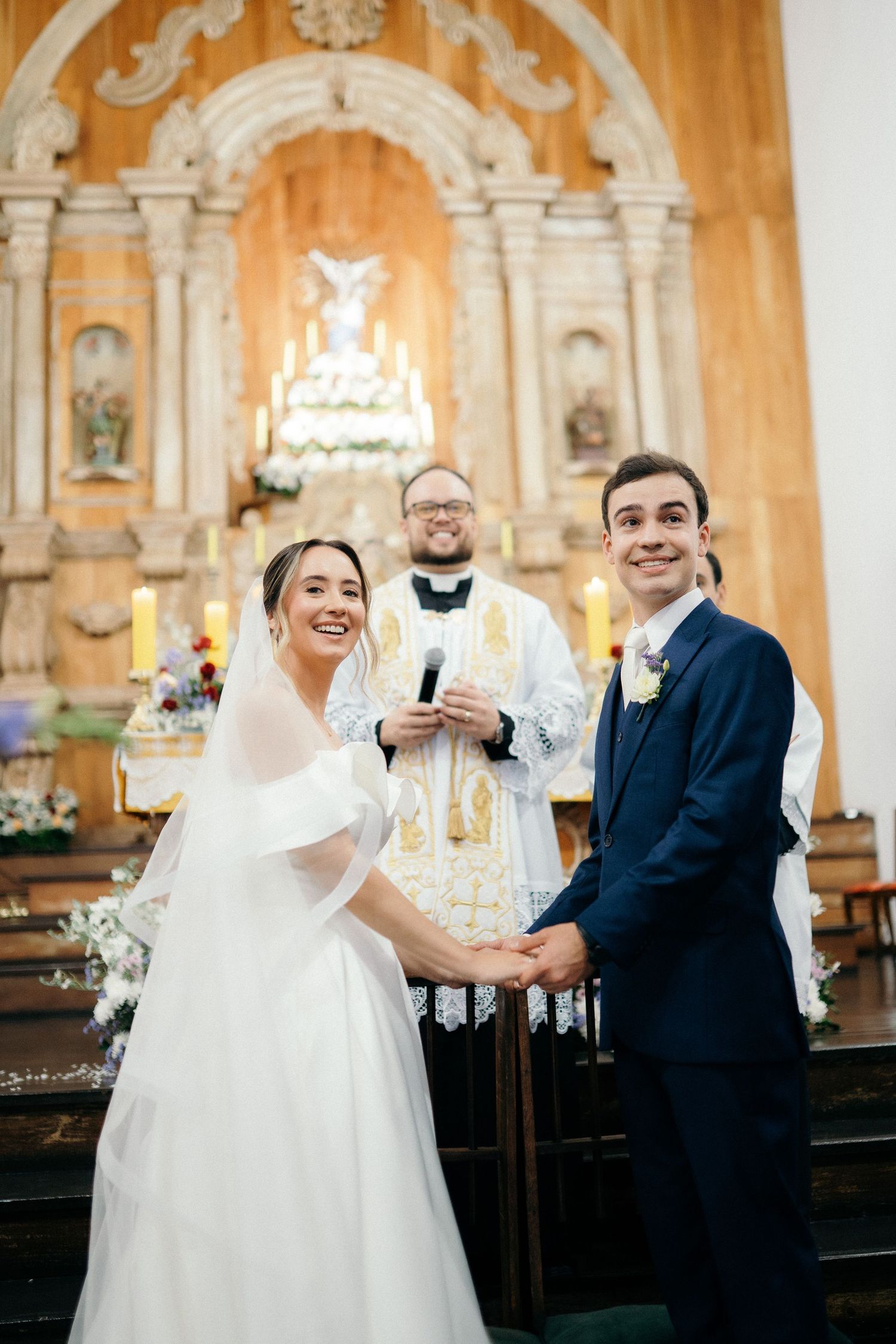Joyful bride and groom holding hands at church altar with priest officiating during Catholic wedding ceremony.
