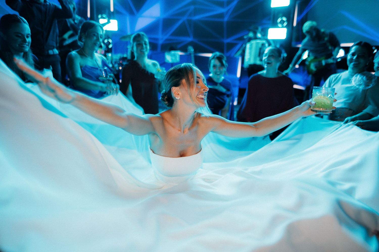 Joyful bride in a flowing white gown dancing with arms outstretched under blue lighting at wedding reception.